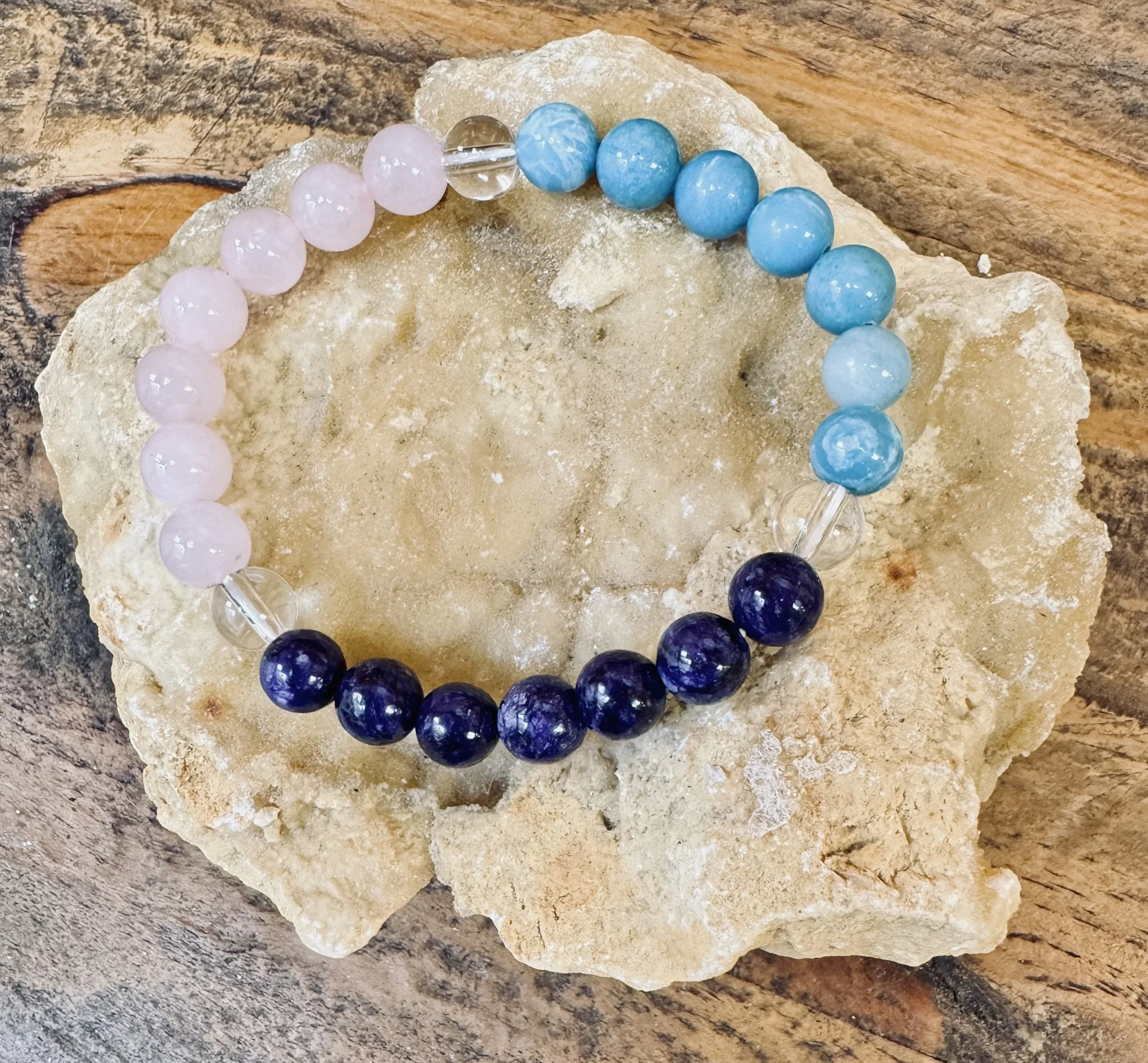 A beaded bracelet with pink, clear, light blue, and dark blue beads resting on a light-colored rock on a wooden surface.