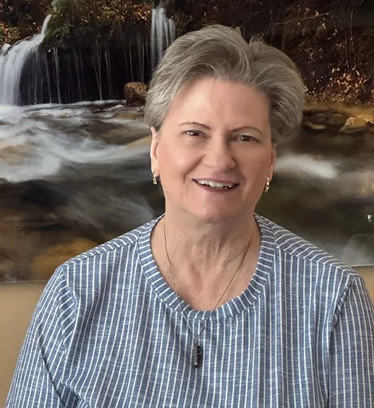 An older woman with short gray hair smiling in front of a waterfall background.