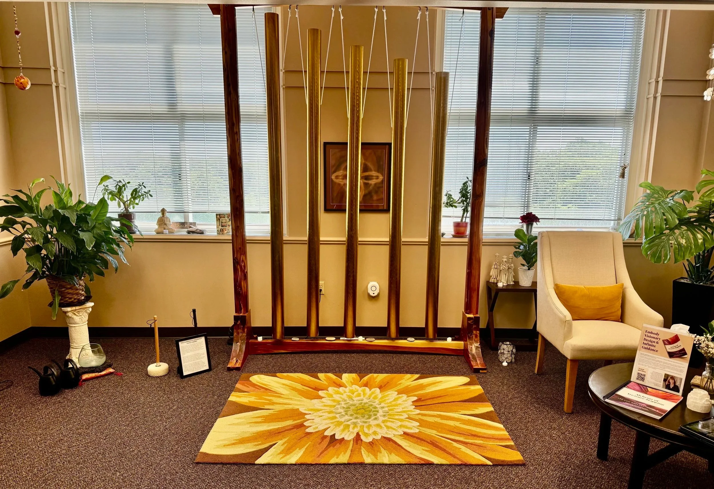 Interior of a room with large windows with blinds, potted plants on either side, a beige armchair with a yellow pillow, a colorful floral rug, and a wooden structure with of Gong Chimes.