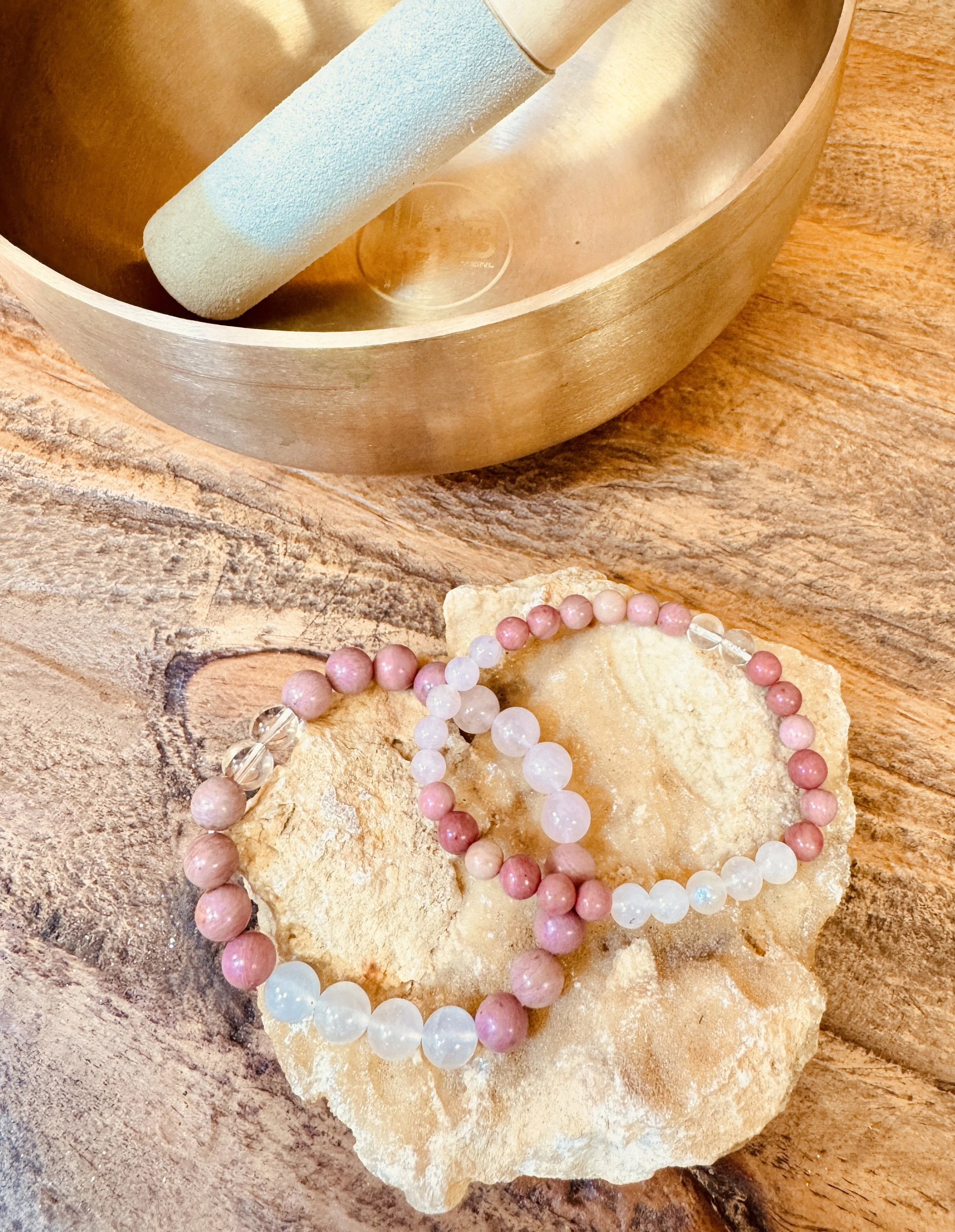 A close-up of a wooden surface with a pink and white beaded necklace resting on a piece of light-colored stone. A brass bowl is visible in the background.