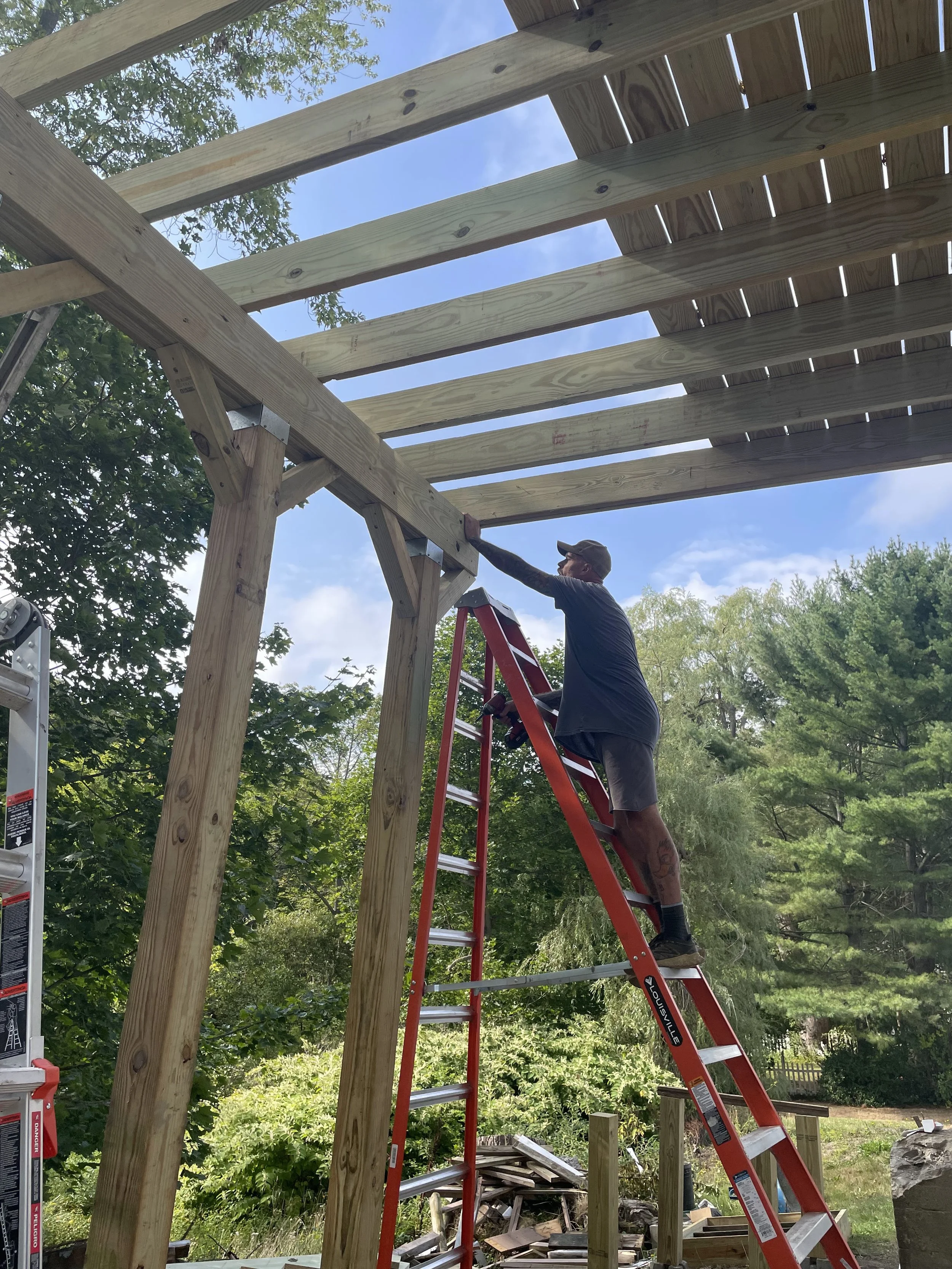 A person on a ladder in a baseball cap, shorts, boots, and blue shirt is reaching toward the underneath corner of a second-story deck. Above them the floor is on its way toward being complete, about half the deck done.