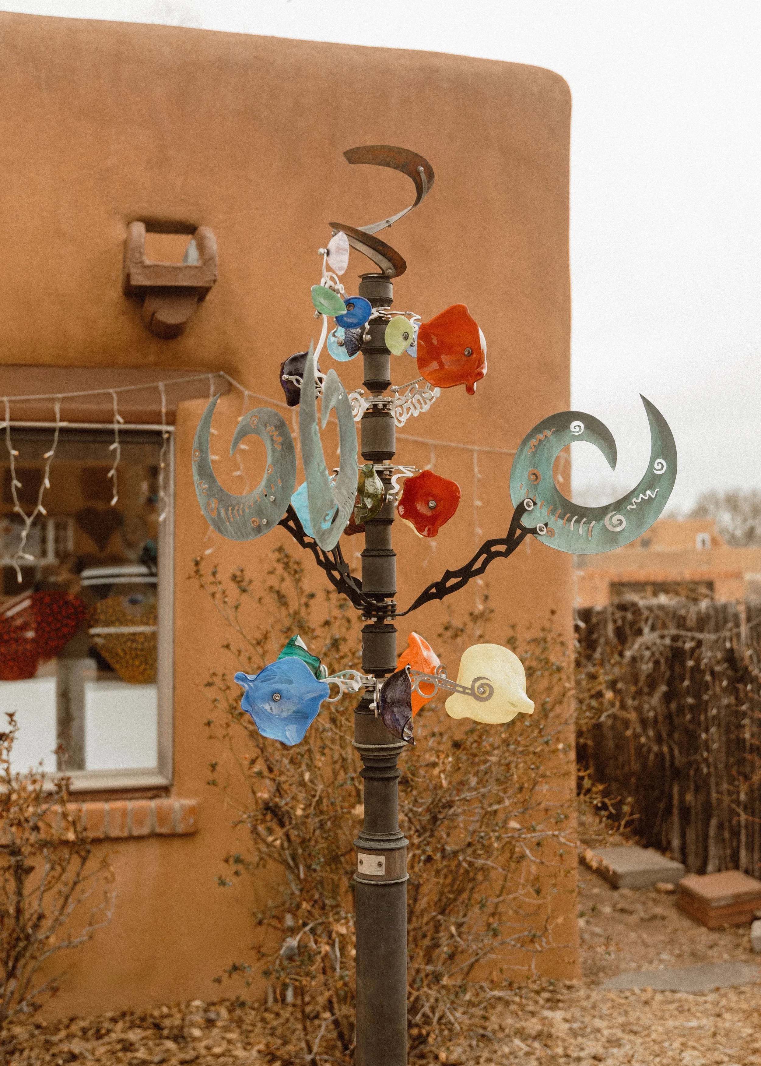 Colorful wind chimes hanging from a black metal pole in front of an adobe-style building and leafless bushes.