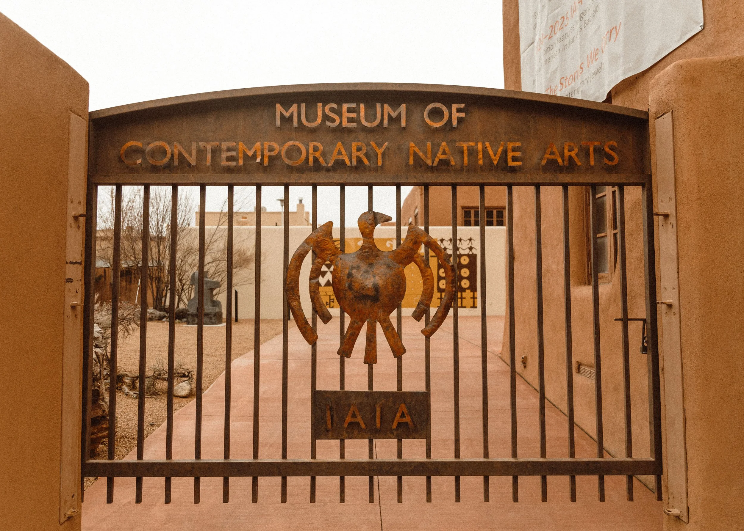 Steel gate with sign for Museum of Contemporary Native Arts, featuring a stylized Native American figure and the IAIA logo, with a building and desert landscaping in the background.