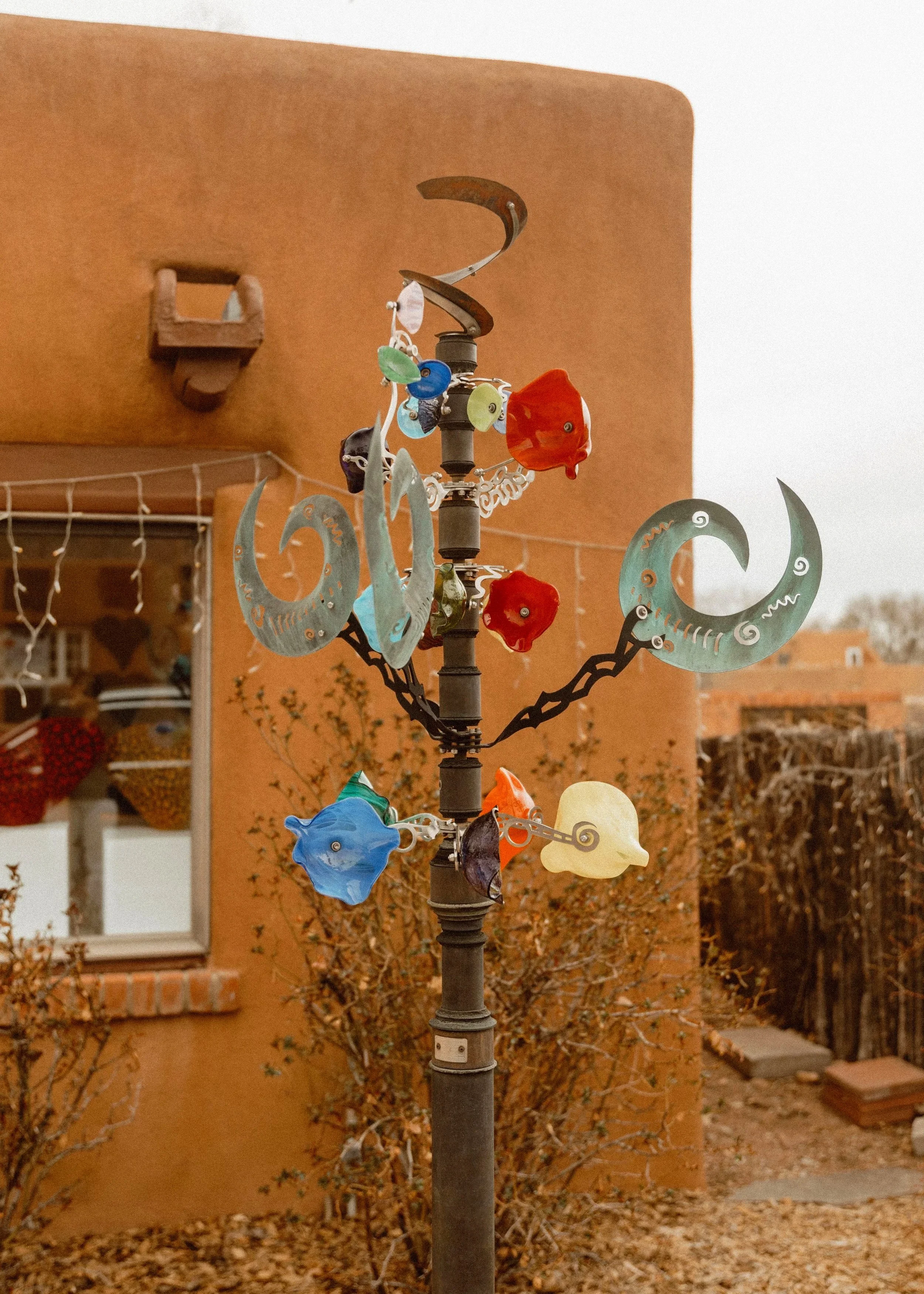Colorful metal wind chimes hanging on a garden post with a brick house and autumn bushes in the background.
