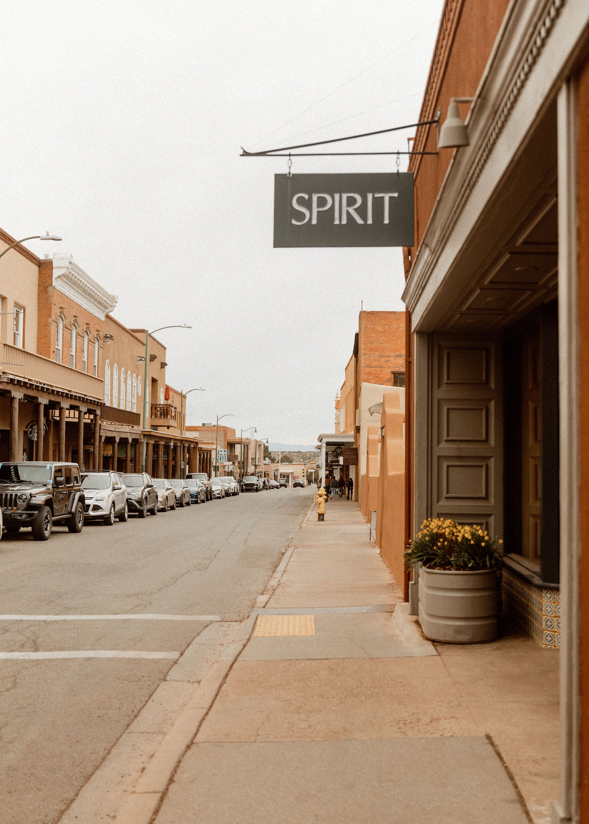 A daytime street view of a small town with parked cars, brick buildings, and a sign hanging from a building that says 'SPIRIT.'