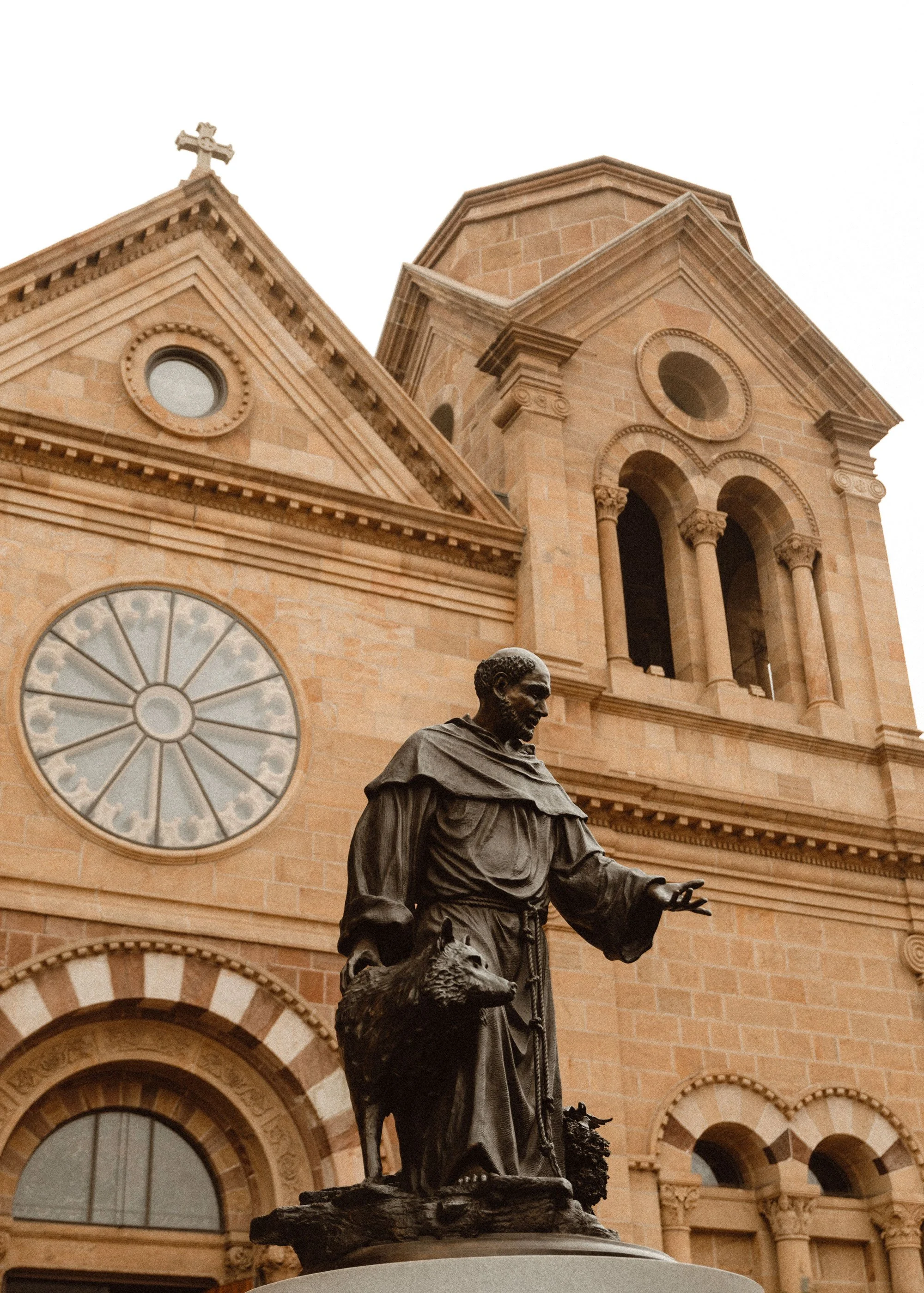 A bronze statue of a man dressed in robes with a wolf at his side, standing in front of a large stone church with arched windows and a clock tower.