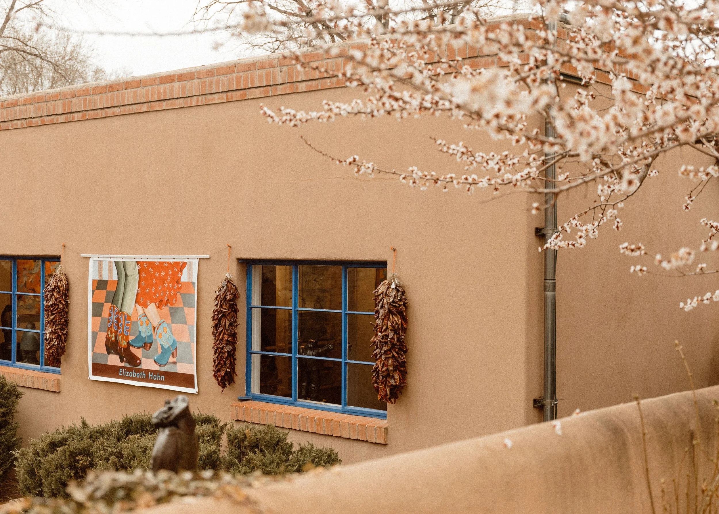 A building with peach-colored walls and blue window frames, decorated with dried herbs hanging on either side of a banner featuring a pair of legs in colorful boots, with the name Elizabeth Hahn below. Cherry blossom branches with pink flowers are vi