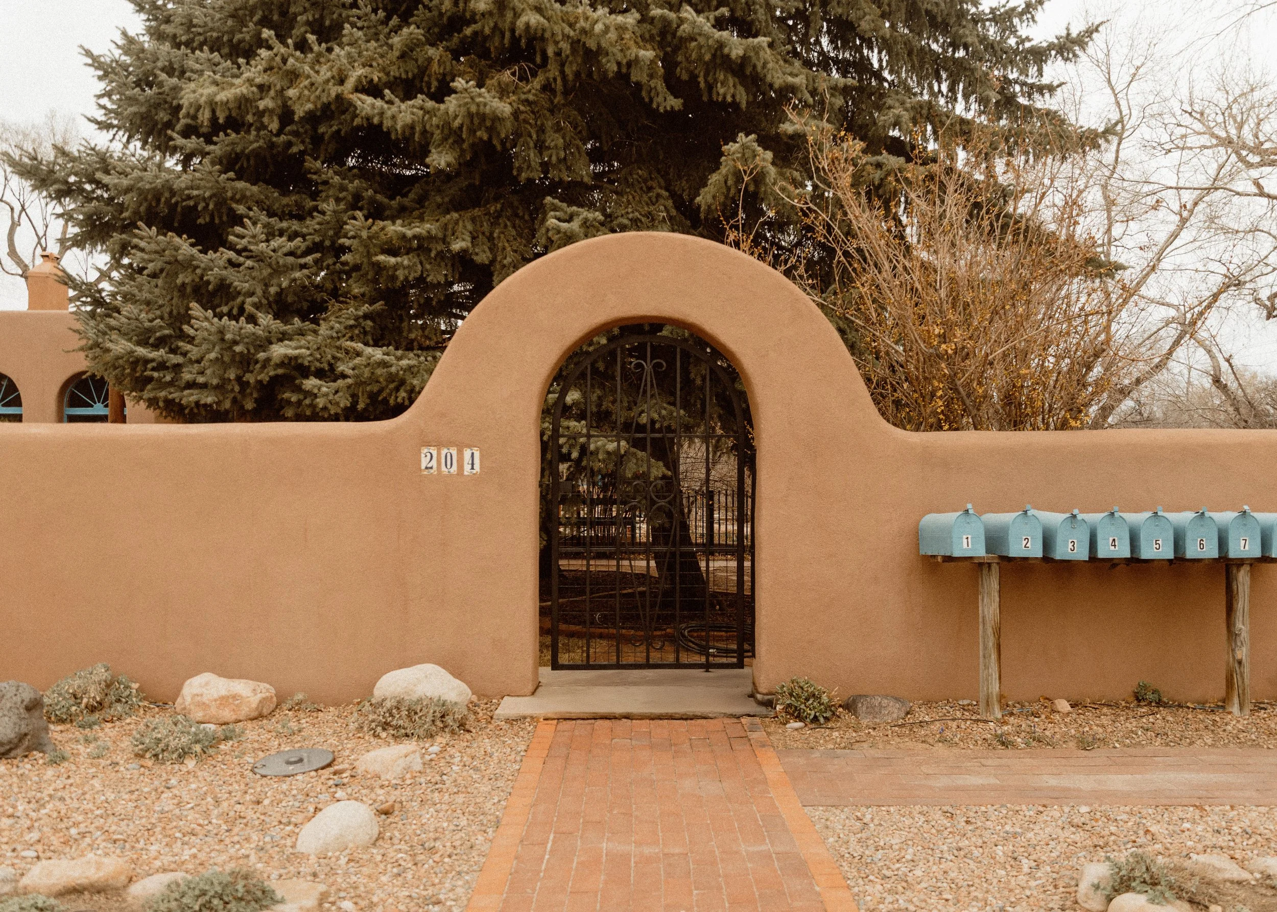 A desert-style stucco wall with a rounded gate, house number 204, metal gate, blue mailboxes numbered 1 to 7 on wooden posts, rocks and drought-tolerant plants in front, large evergreen tree, and leafless trees in the background.