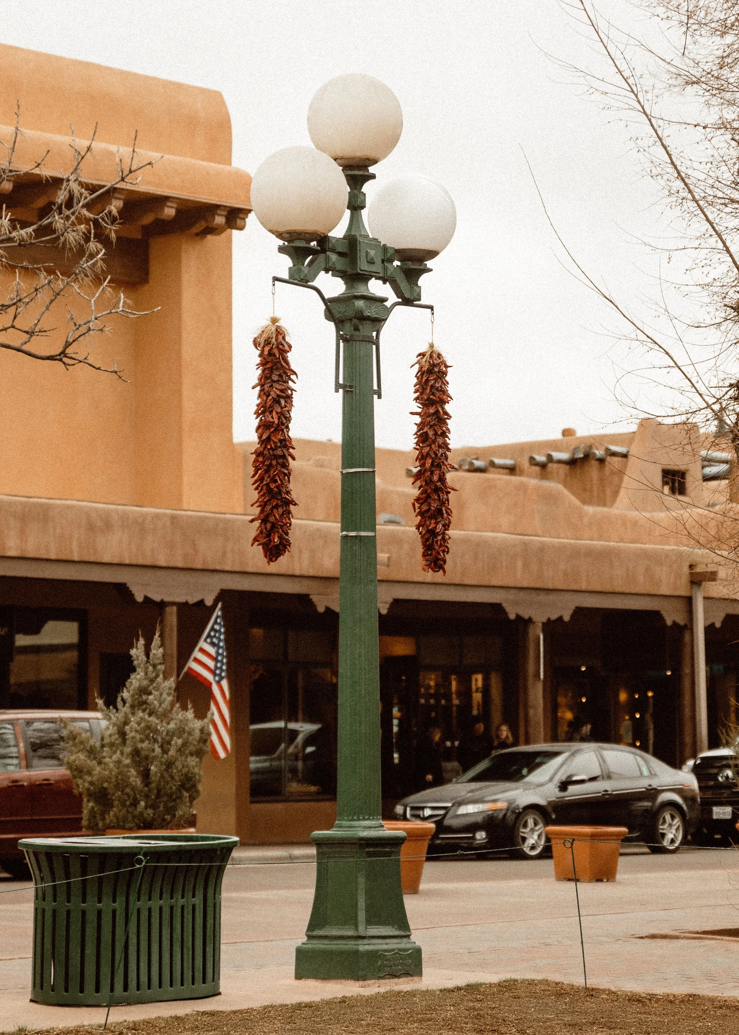 A vintage-style green street lamp with three round white globes, decorated with dried chili pepper strings hanging from the top, located in a small town or city square. There is a red car, an American flag, a trash bin, and some trees and buildings i