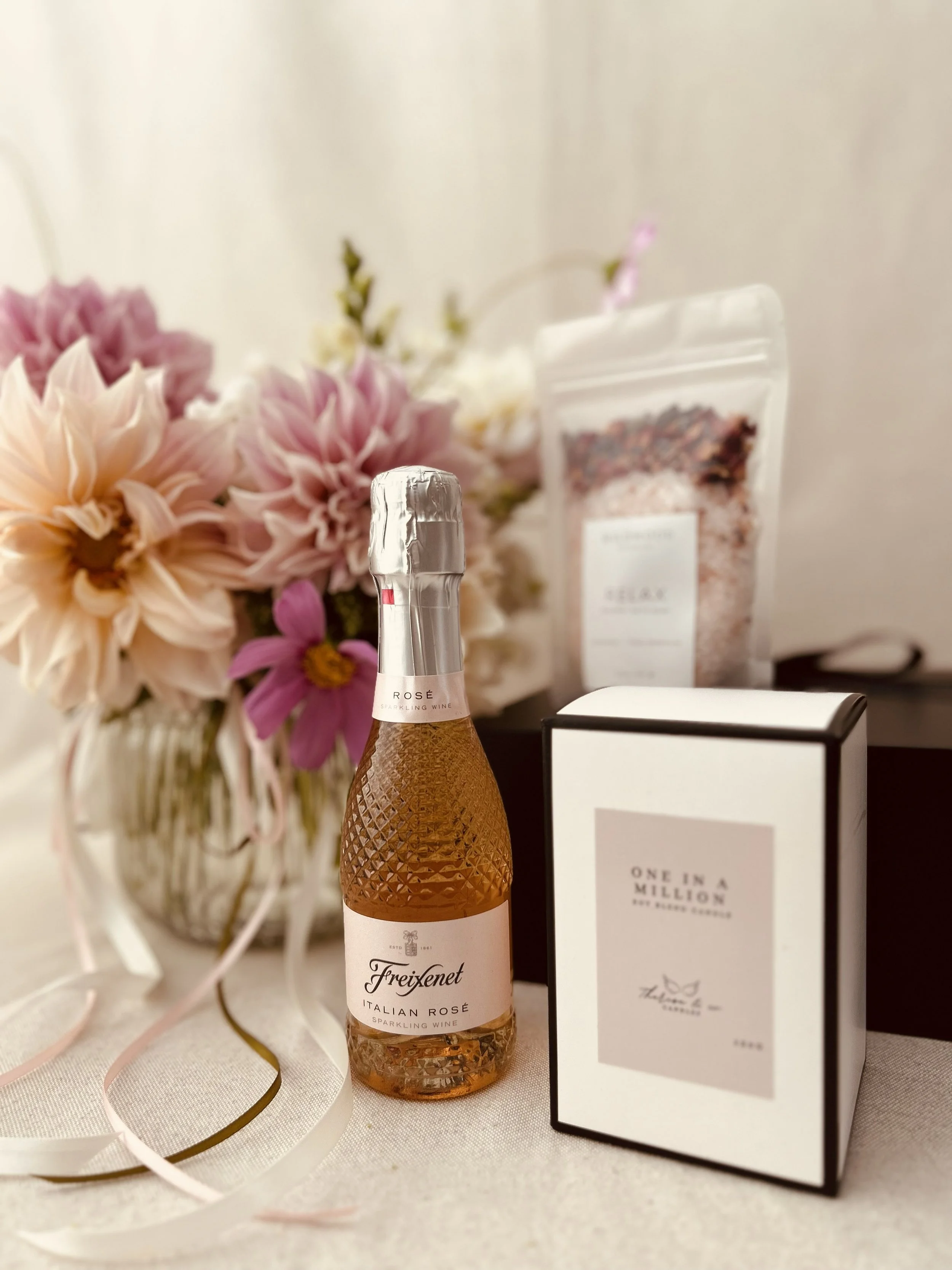 A bottle of Freixenet Italian Rosé sparkling wine on a table with a floral bouquet, a box labeled 'One in a Million,' and a bag of Himalayan salt in the background.