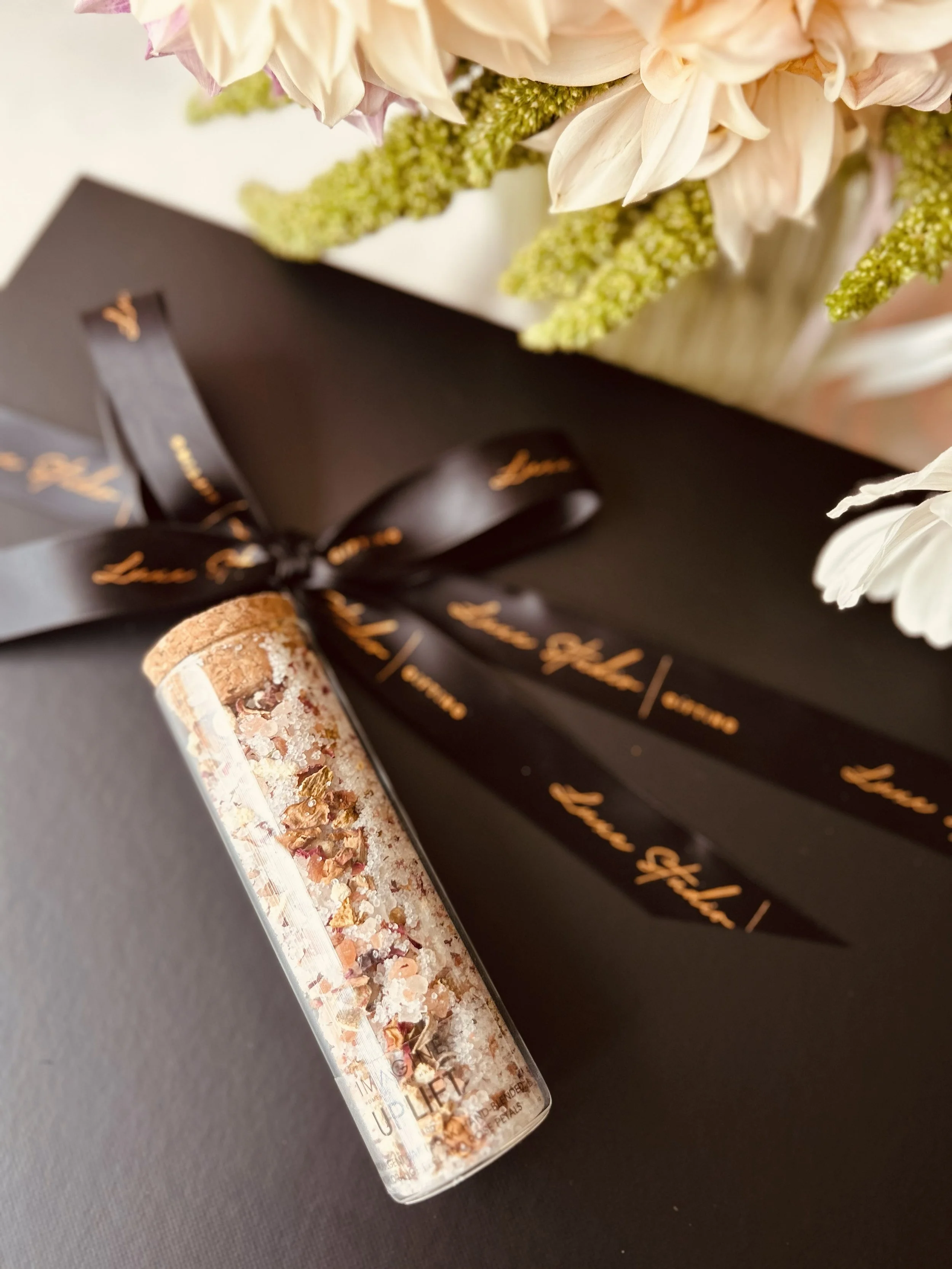 A black gift box with a black ribbon and gold writing, a glass jar with a cork lid partially filled with crushed makeup or bath salts, and a bouquet of white and pale pink flowers.
