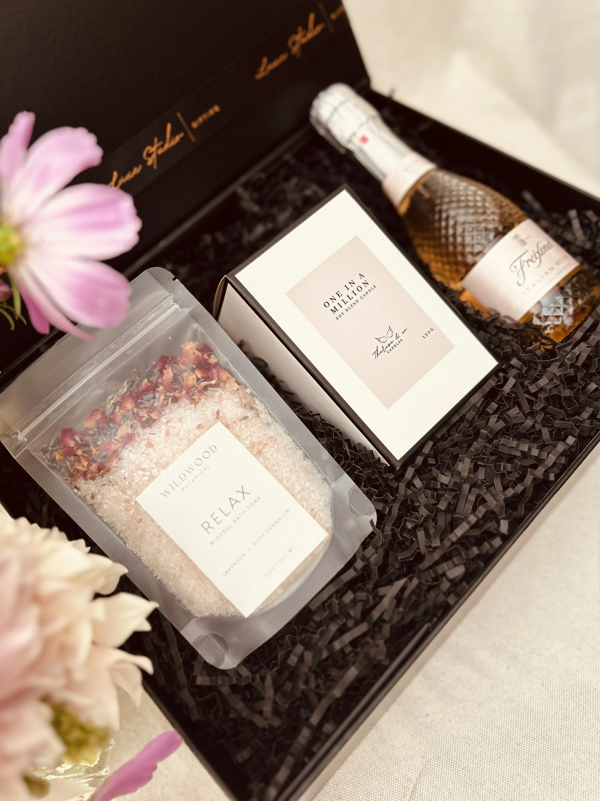 A gift box containing a bag of bath salts labeled 'RELAX', a small box of soy blend candle titled 'ONE IN A MILLION', and a small bottle of Italian sparkling wine 'Freixenet' with a white cap and textured bottle, arranged on black shredded paper inside a gift box.