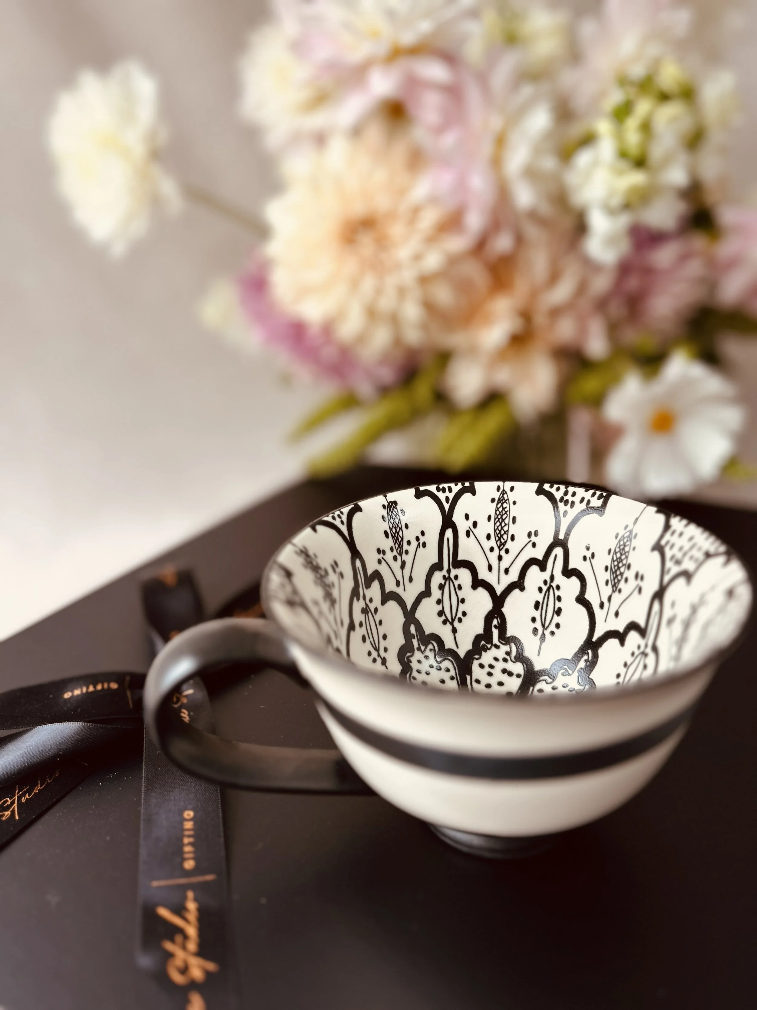 A black and white patterned ceramic mug with floral and leaf designs, resting on a black surface with a black ribbon, and a blurred background of pink and white flowers.