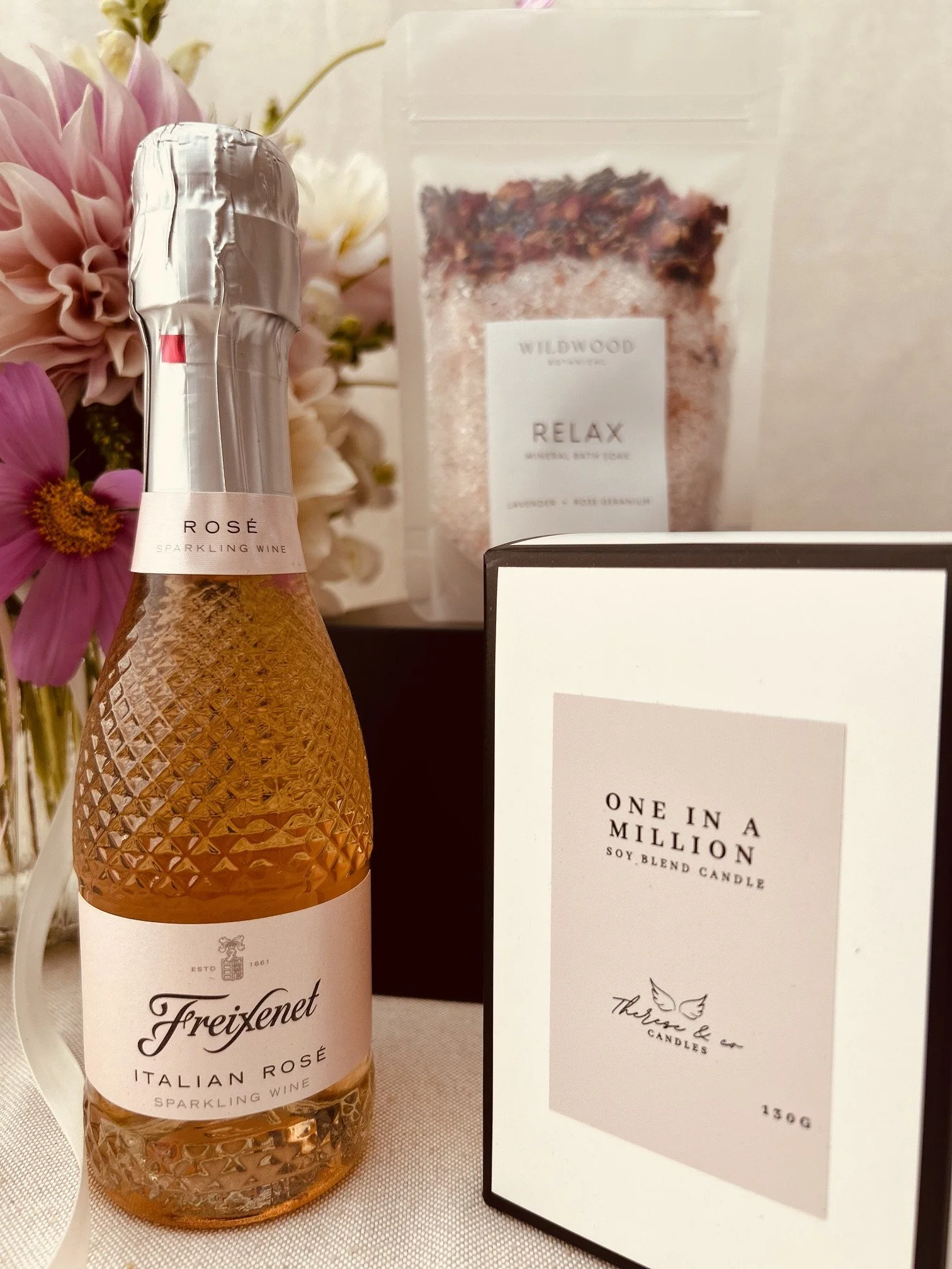 A bottle of Freixenet Italian Rosé sparkling wine, a box of One in a Million soy-blend candle, and a bag of Wildwood Relax mineral bath soak with flowers in the background.