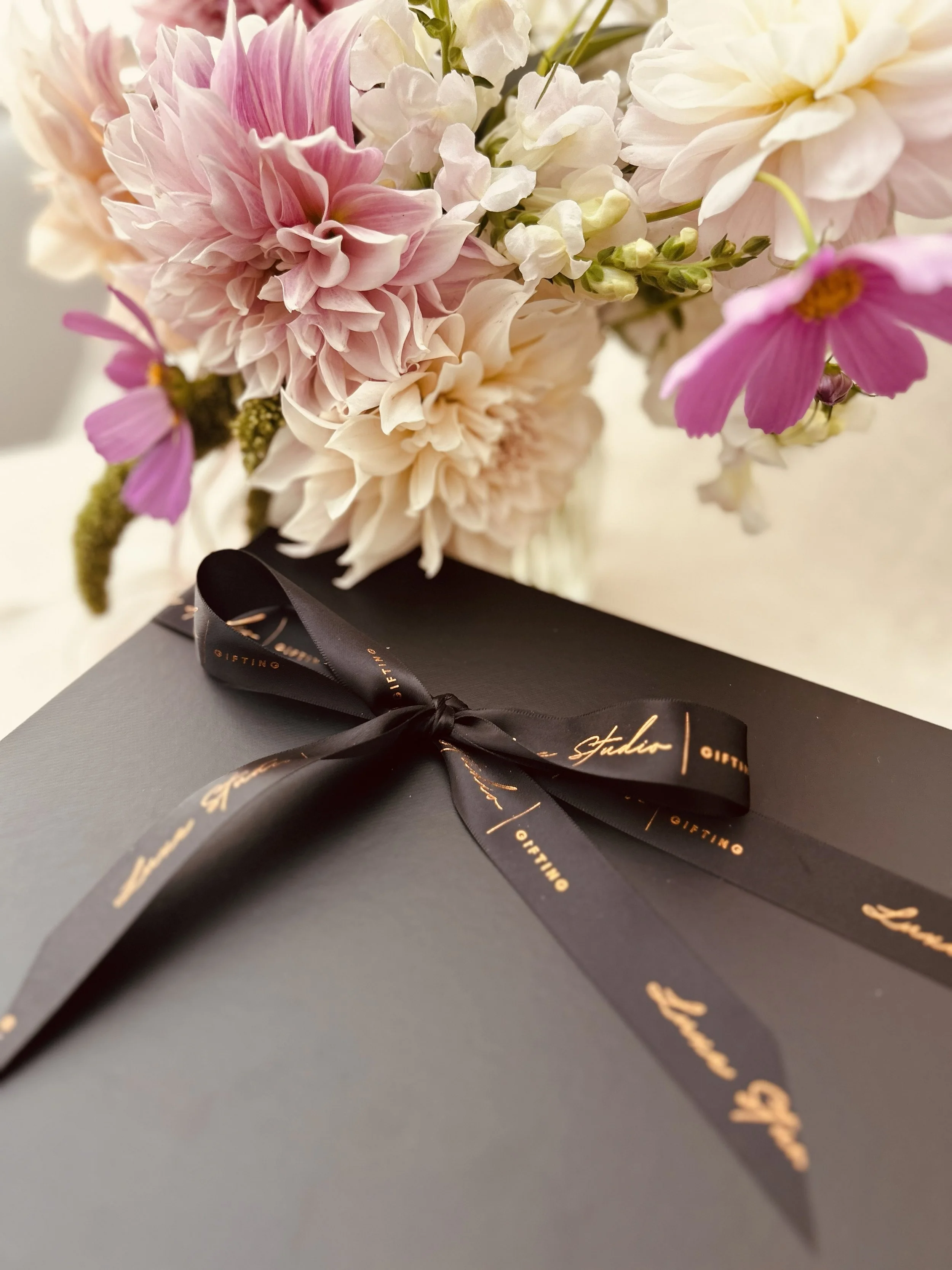 A bouquet of pink and white flowers placed behind a black gift box wrapped with a black ribbon.