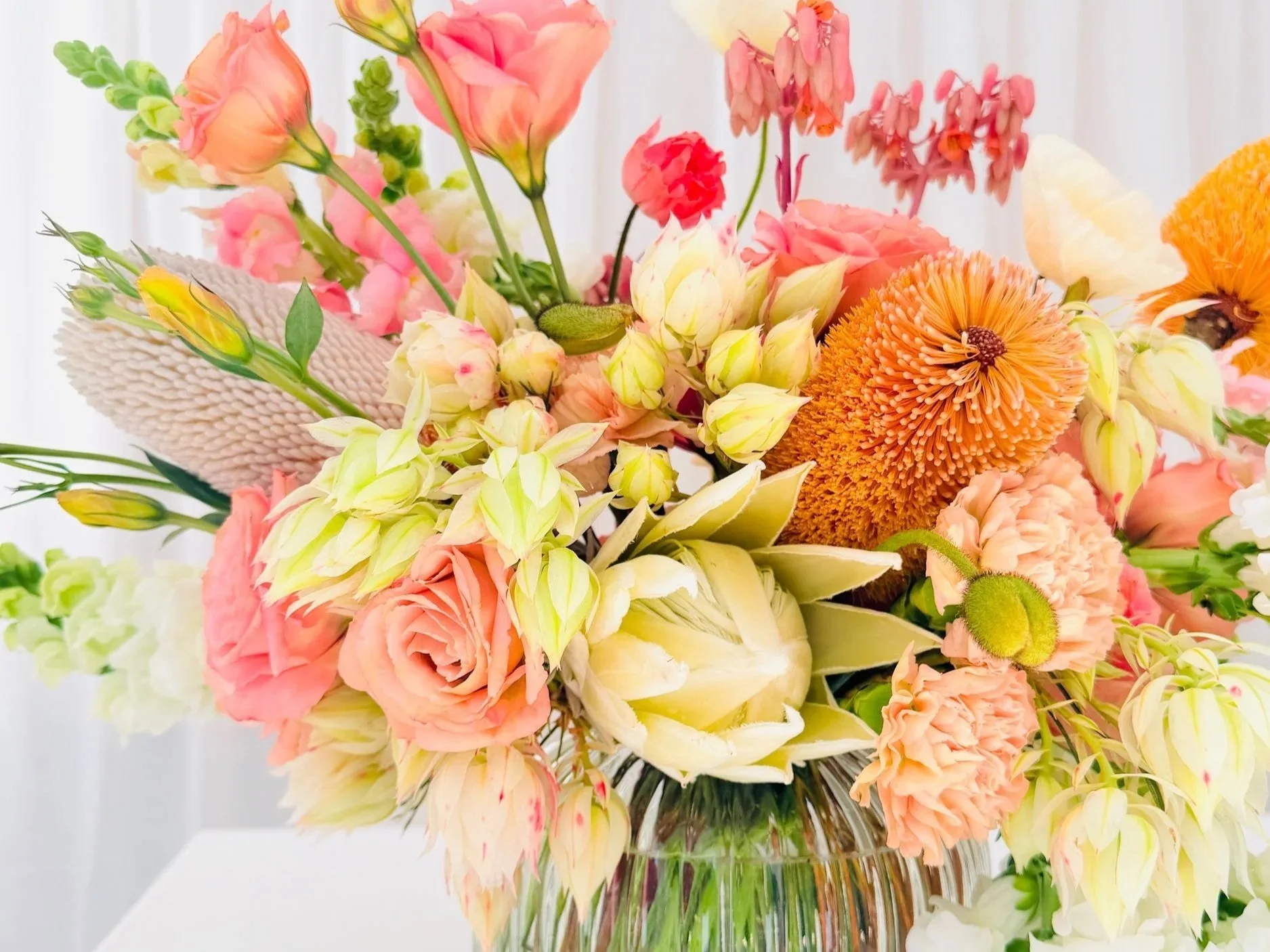 A colorful flower bouquet in a glass vase with pink, orange, and yellow flowers, including roses, lilies, and other blossoms, set against a light background.