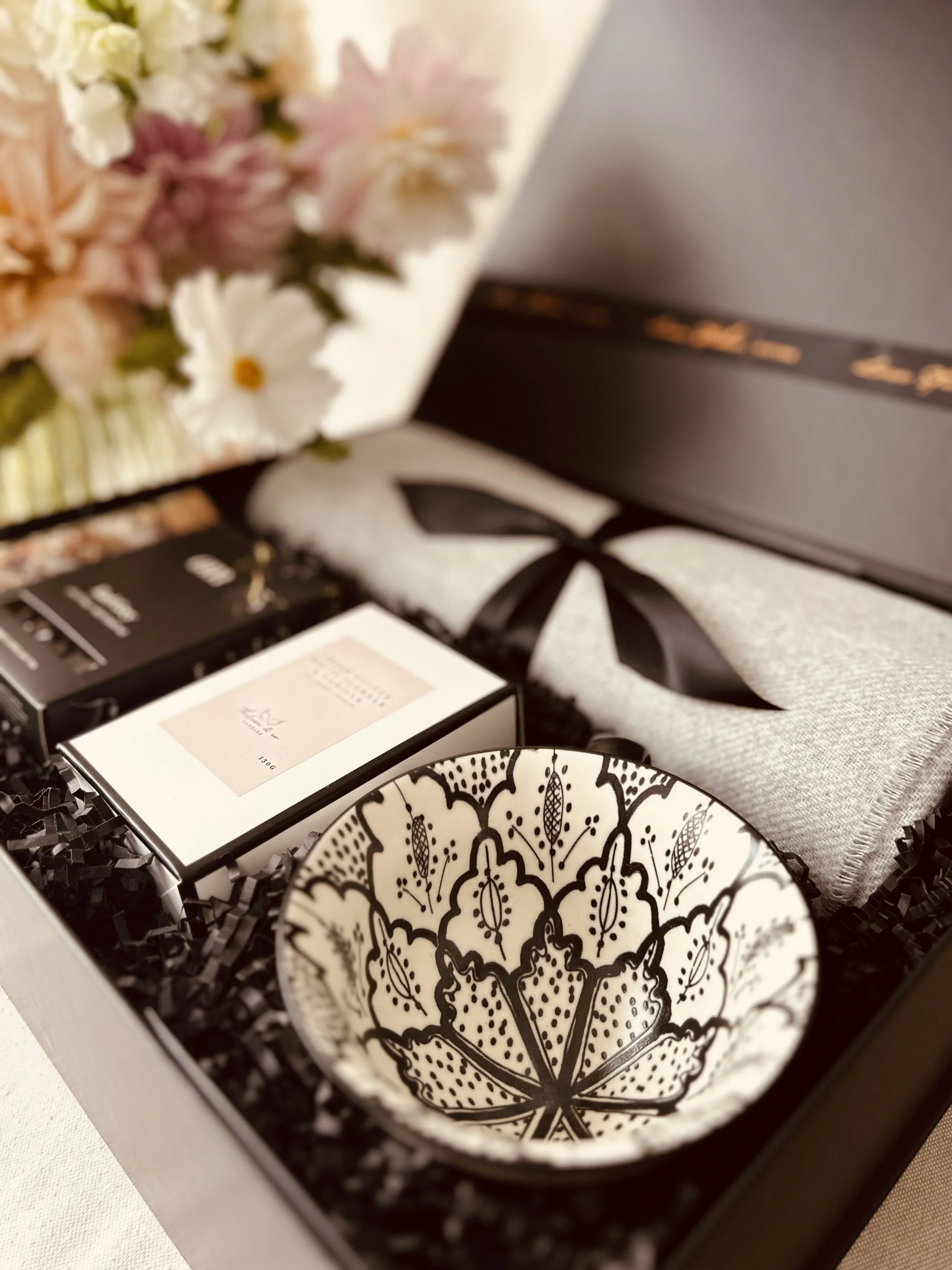 Gift box with skincare products, a decorative bowl, and a towel with a black ribbon, accompanied by a flower bouquet in the background.