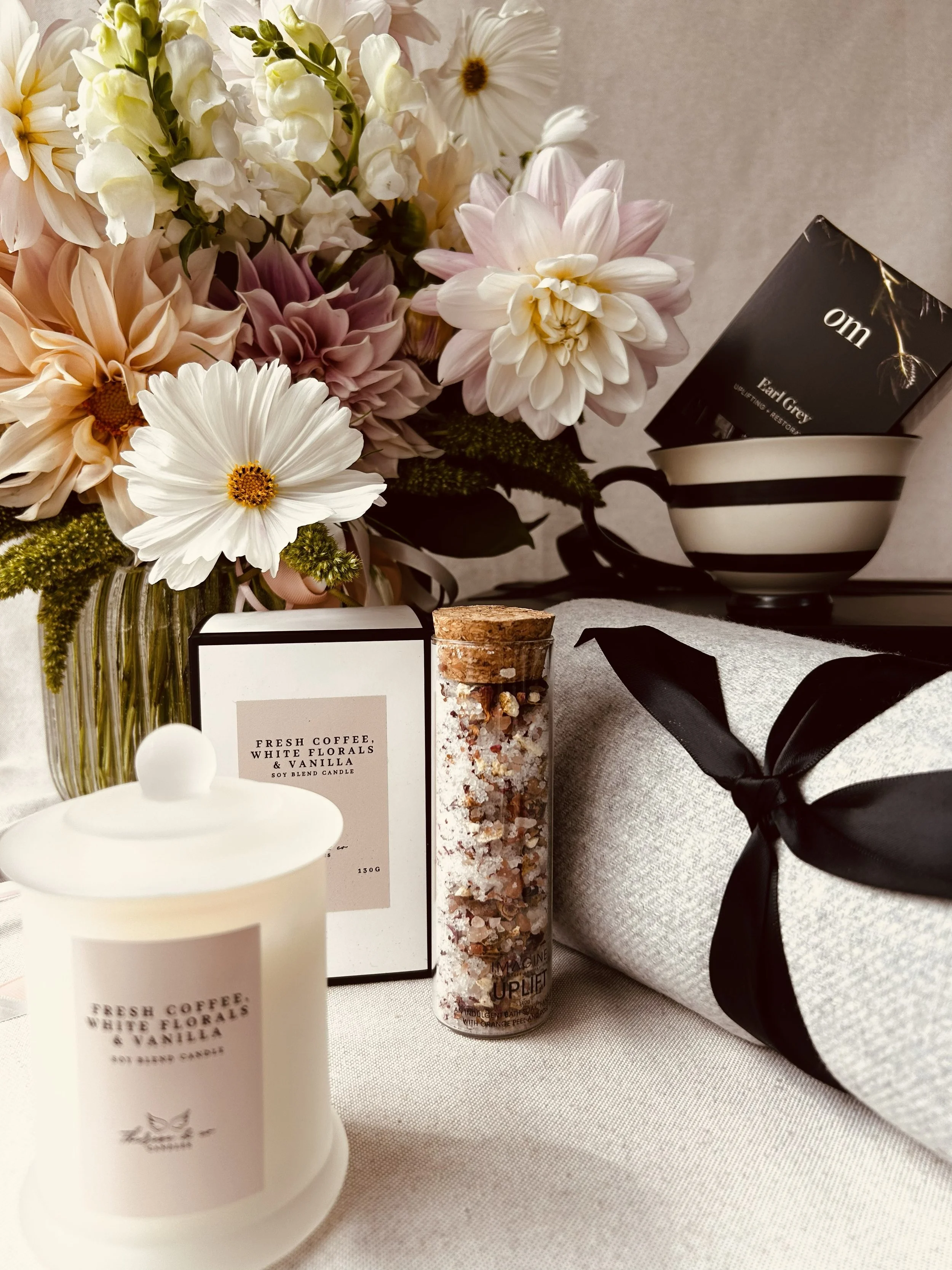 A floral arrangement with white and pink flowers, a candle labeled "Fresh Coffee, White Florals & Vanilla," a glass jar of bath salts, a wrapped gift with a black ribbon, and a black and white striped bowl containing a black box labeled "om" with "Pearl Grey" tea inside.