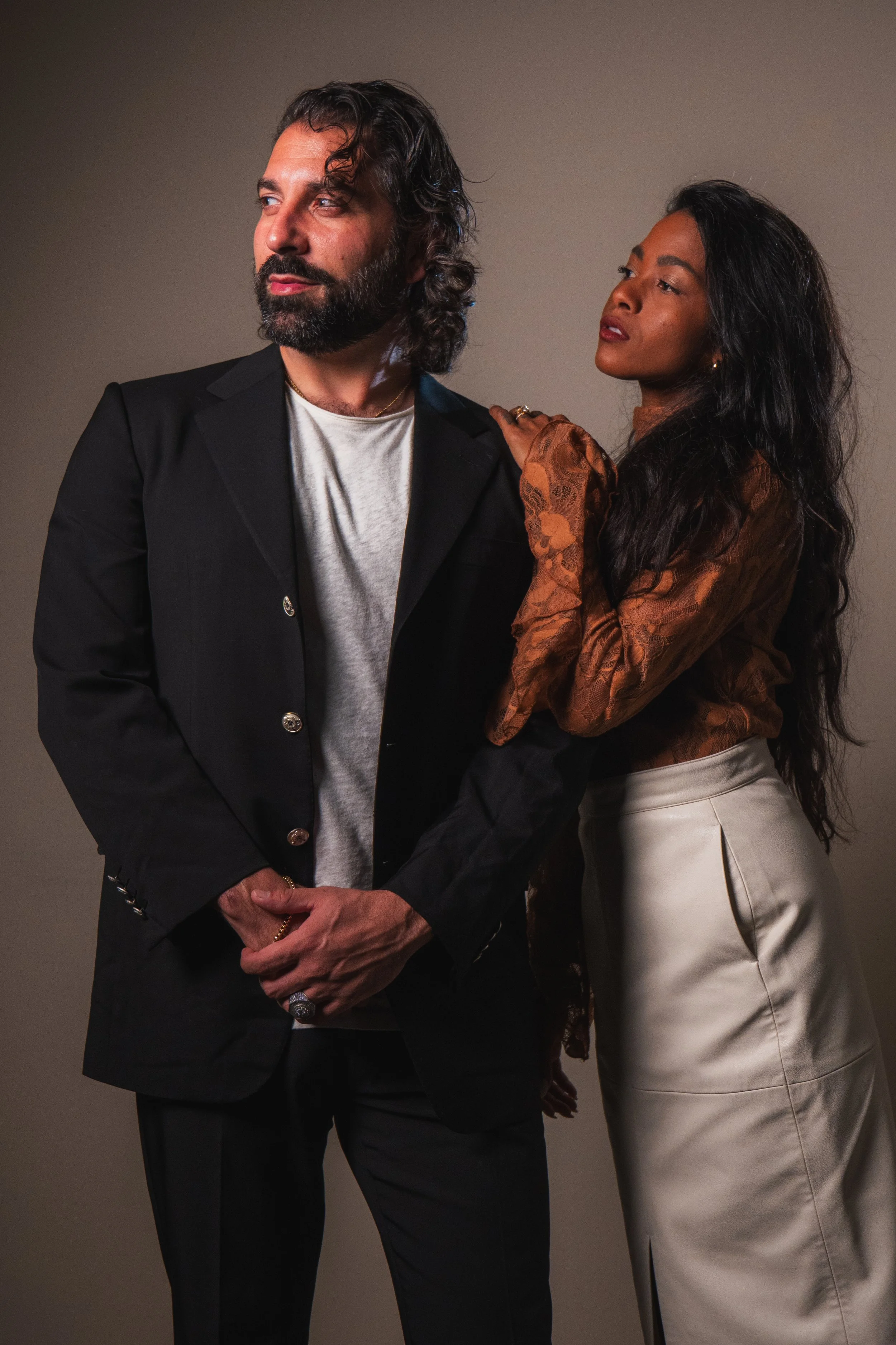 A man and woman posing together, the man has shoulder-length grayish curly hair, beard, and is wearing a black blazer and t-shirt, while the woman has long dark wavy hair, wearing a brown lace blouse and a cream-colored skirt.