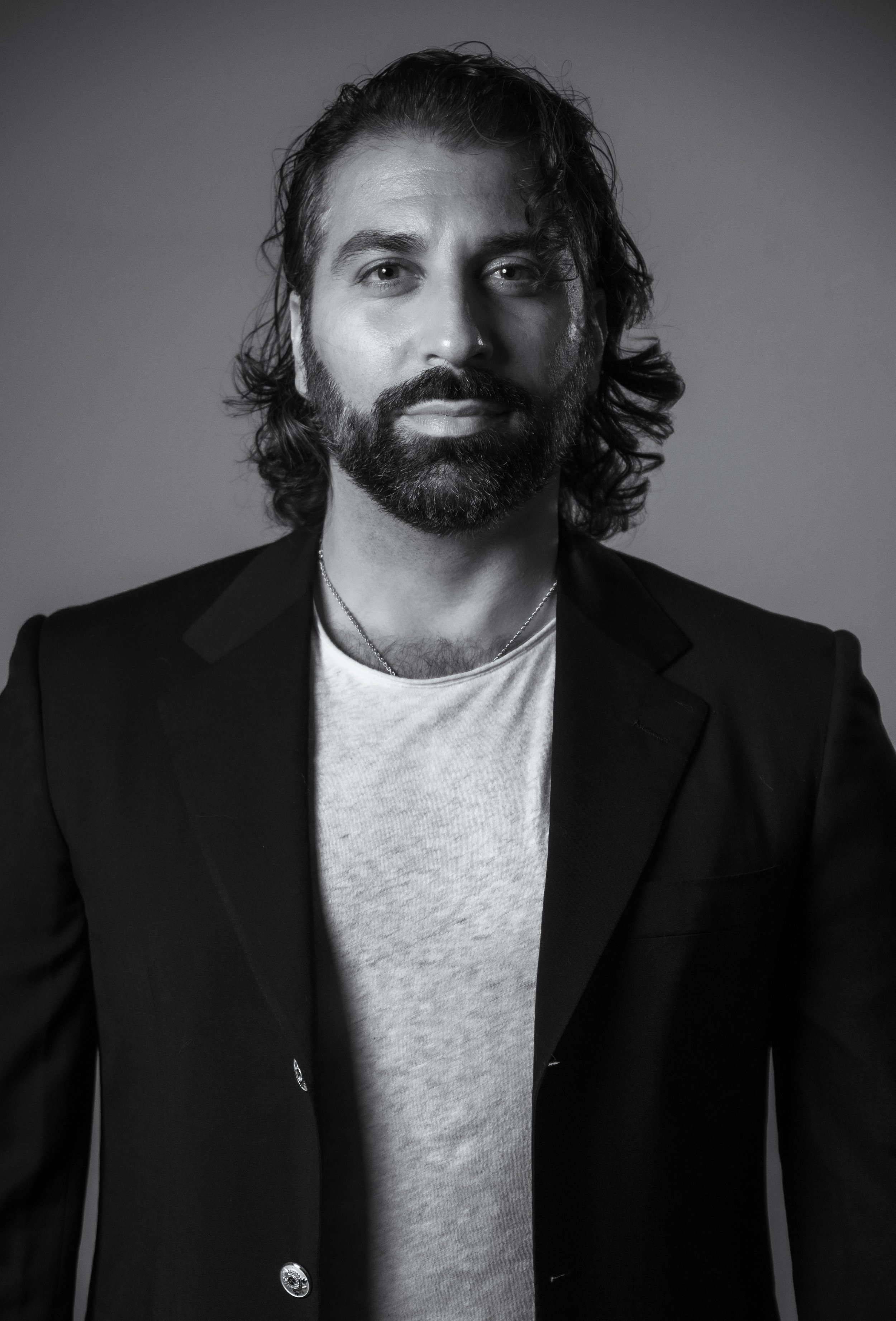 Black and white portrait of a man with wavy hair and a beard, wearing a blazer and a t-shirt, looking directly at the camera.