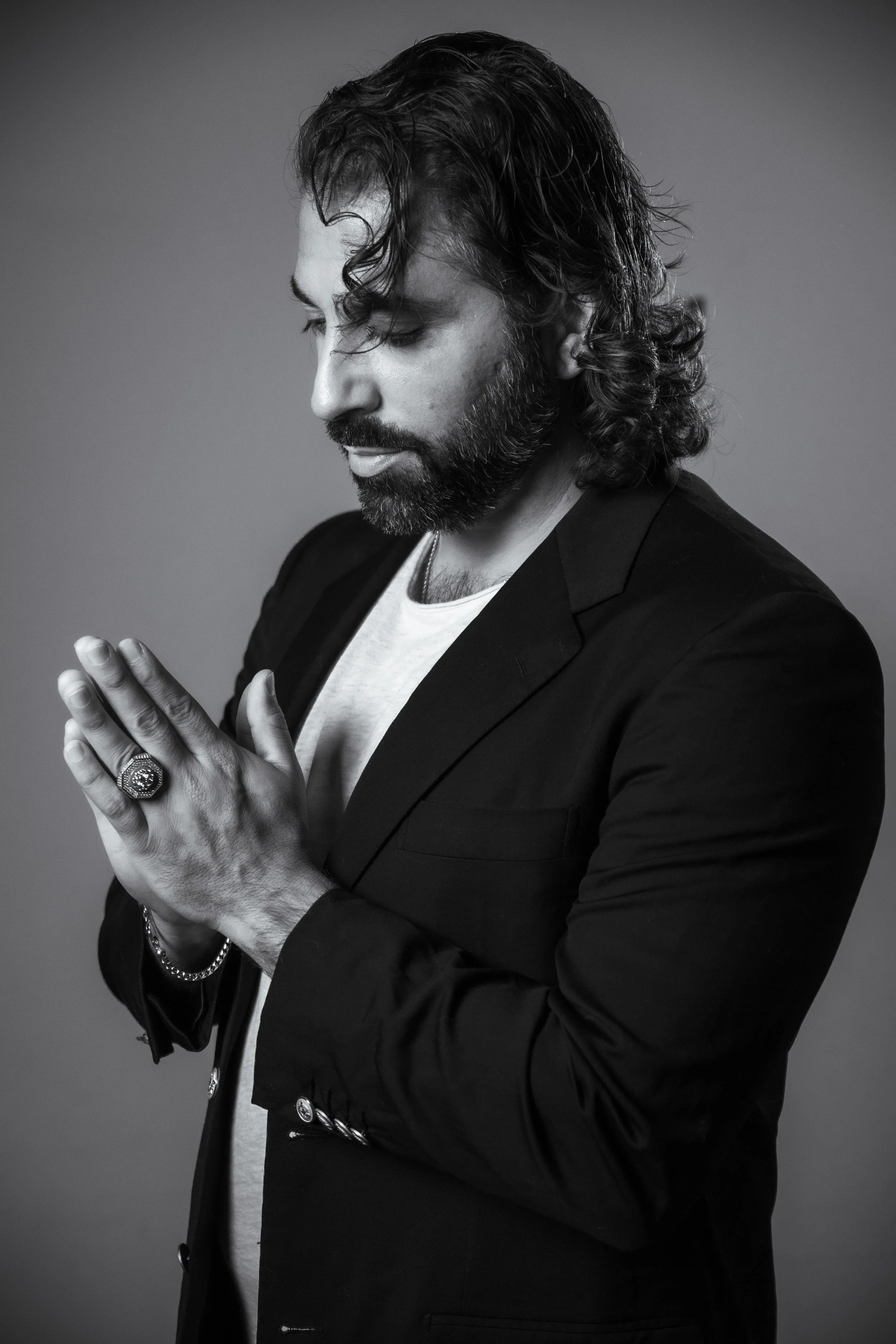 Black and white portrait of a man with long wavy hair and a beard, wearing a dark blazer and a white shirt, with his hands pressed together in a prayer-like gesture, looking down.