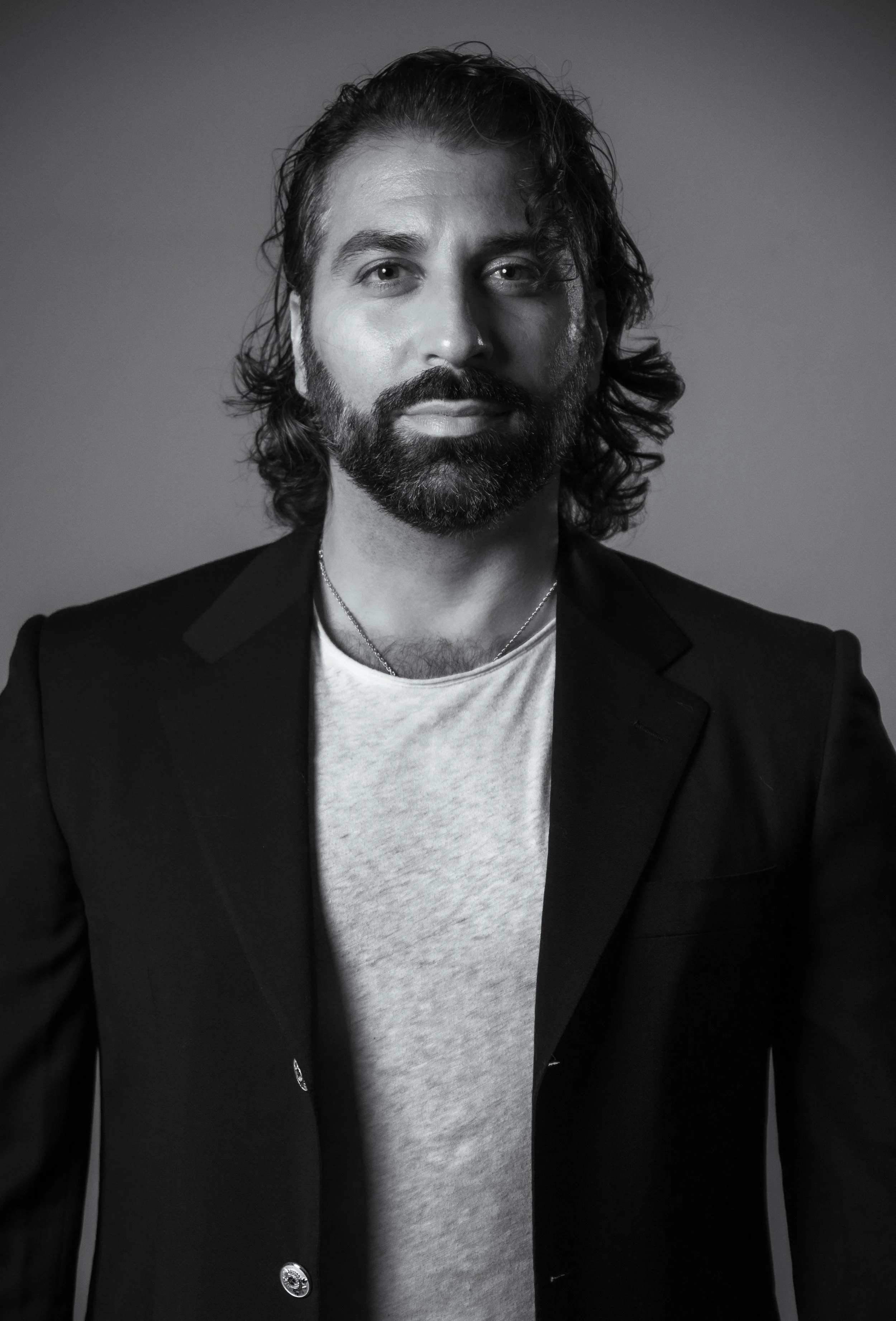 Black and white portrait of a man with long wavy hair and a beard, wearing a black blazer over a light-colored T-shirt, looking directly at the camera.