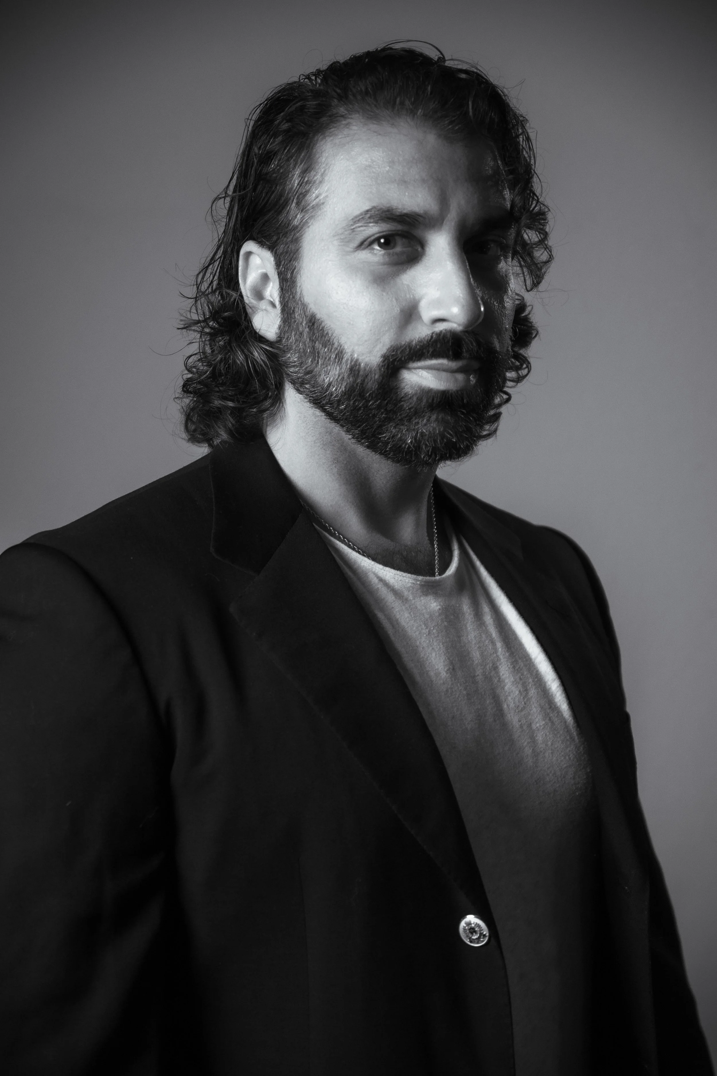 Black and white portrait of a man with long, wavy hair and a beard, dressed in a blazer and t-shirt, looking at the camera.