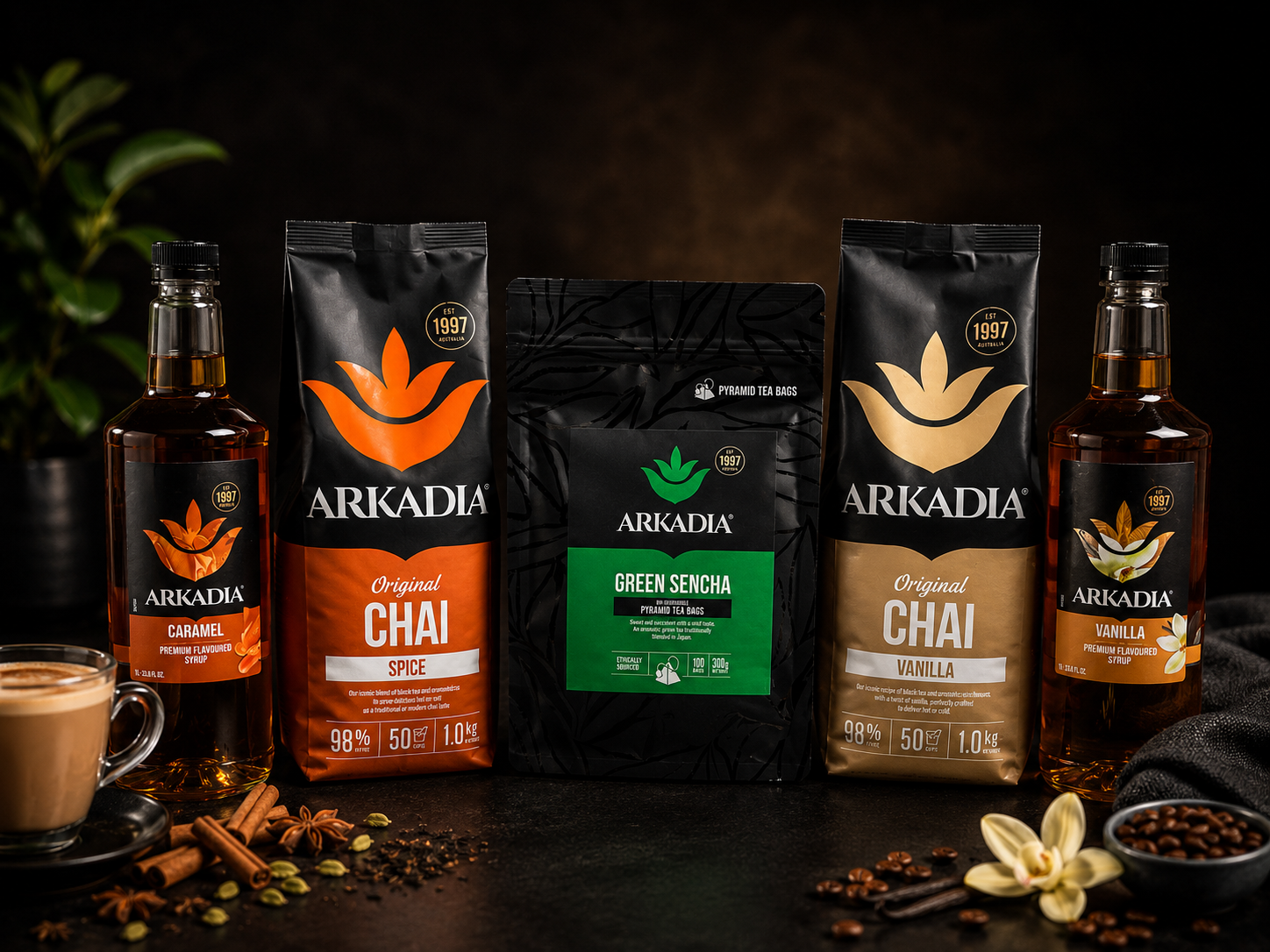 Arrangement of Arkadia chai and tea products, including two bottles of flavored syrup, three packages of chai and green sencha tea bags, surrounded by spices and a cup of chai, on a dark surface with plants in the background.