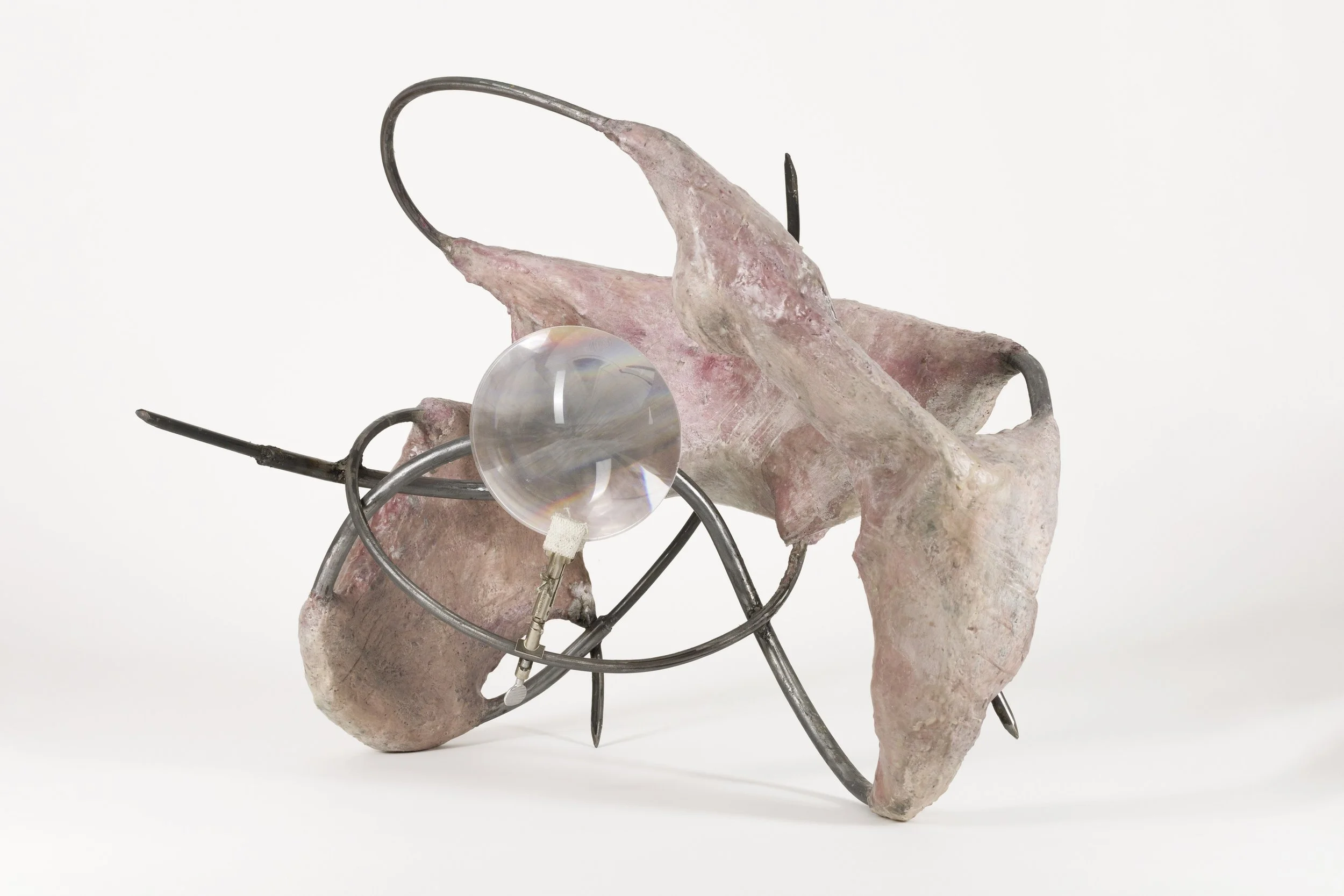 An abstract sculpture featuring pinkish rock, metal wire, a glass disc, and black metal elements against a plain white background.
