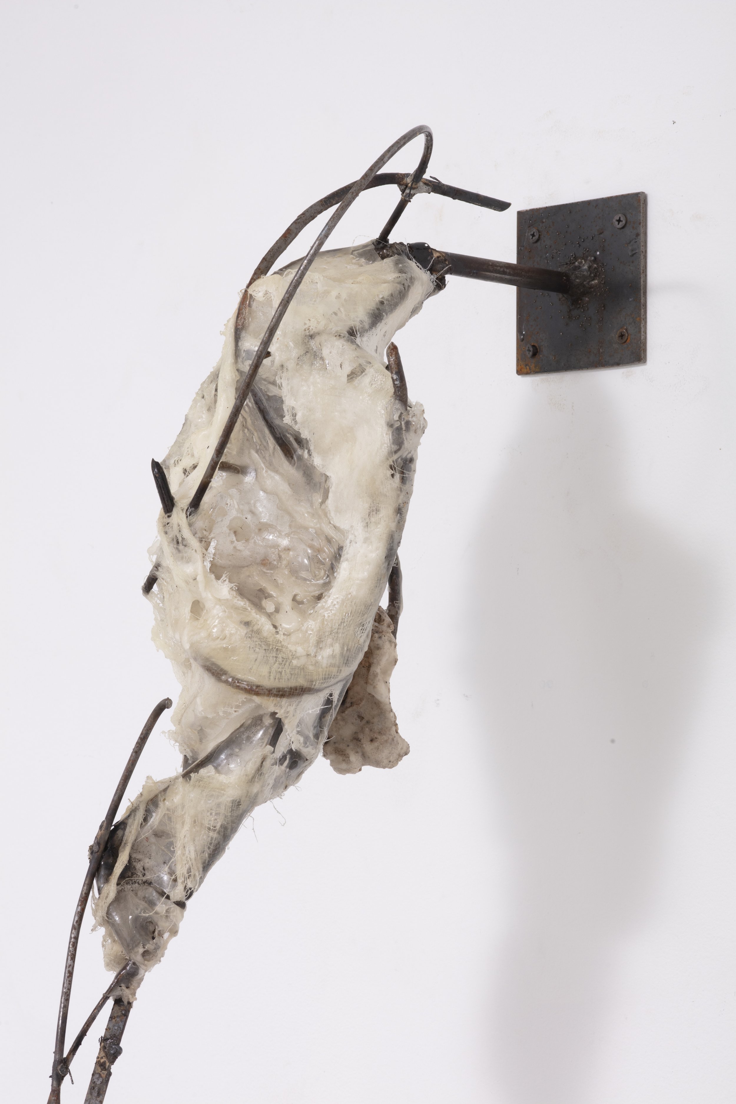 An abstract sculpture composed of intertwined metal rods, with a large irregularly shaped white material resembling resin or wax, mounted on a plain white wall.