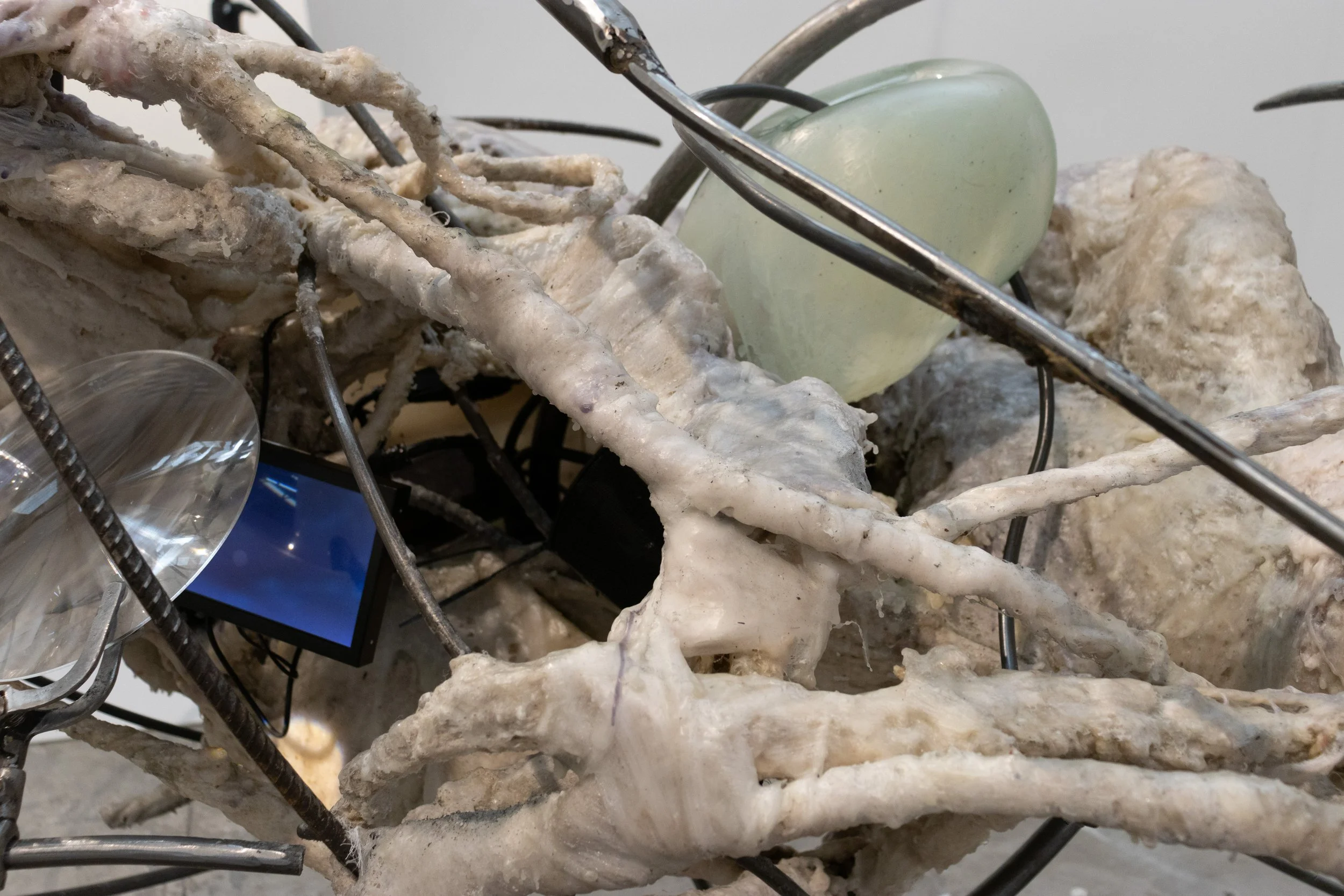 Close-up of a metal wire sculpture resembling intertwined bone-like shapes, with a large rock and a light-green glass object in the background.