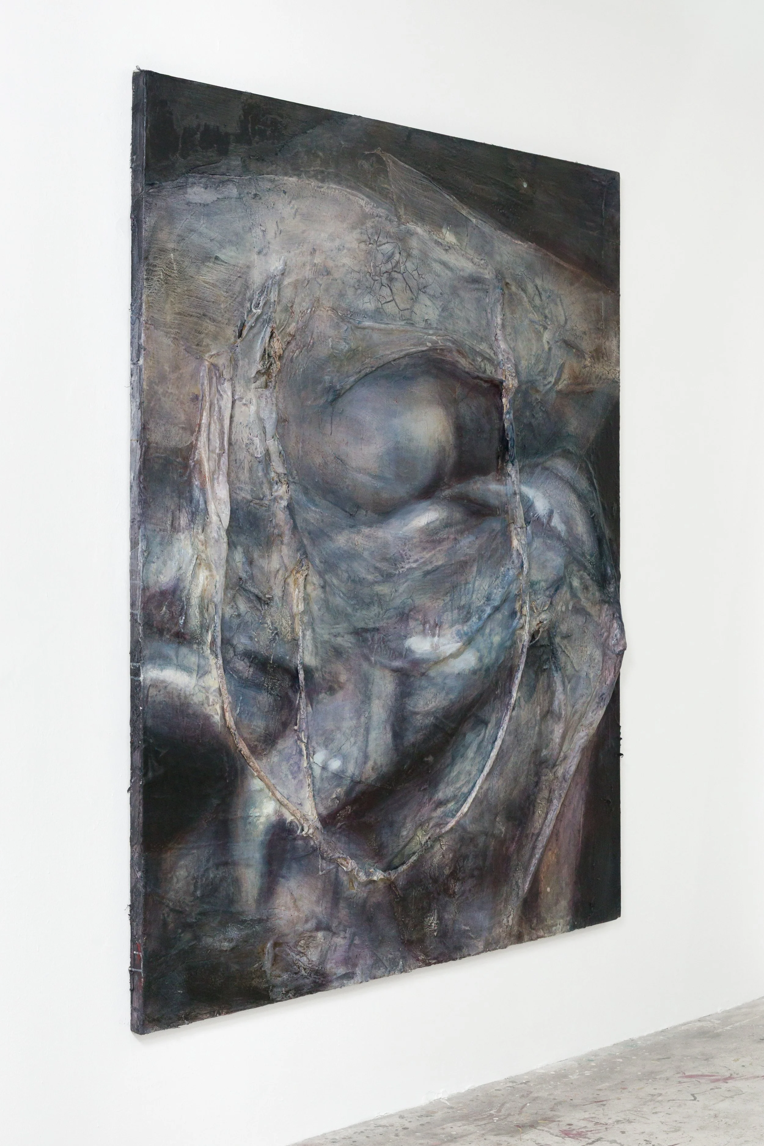 An abstract painting on a large canvas depicts a human face with closed eyes and a large circular element in the forehead, featuring a dark and muted color palette with shades of gray, black, and brown, displayed on a white gallery wall.