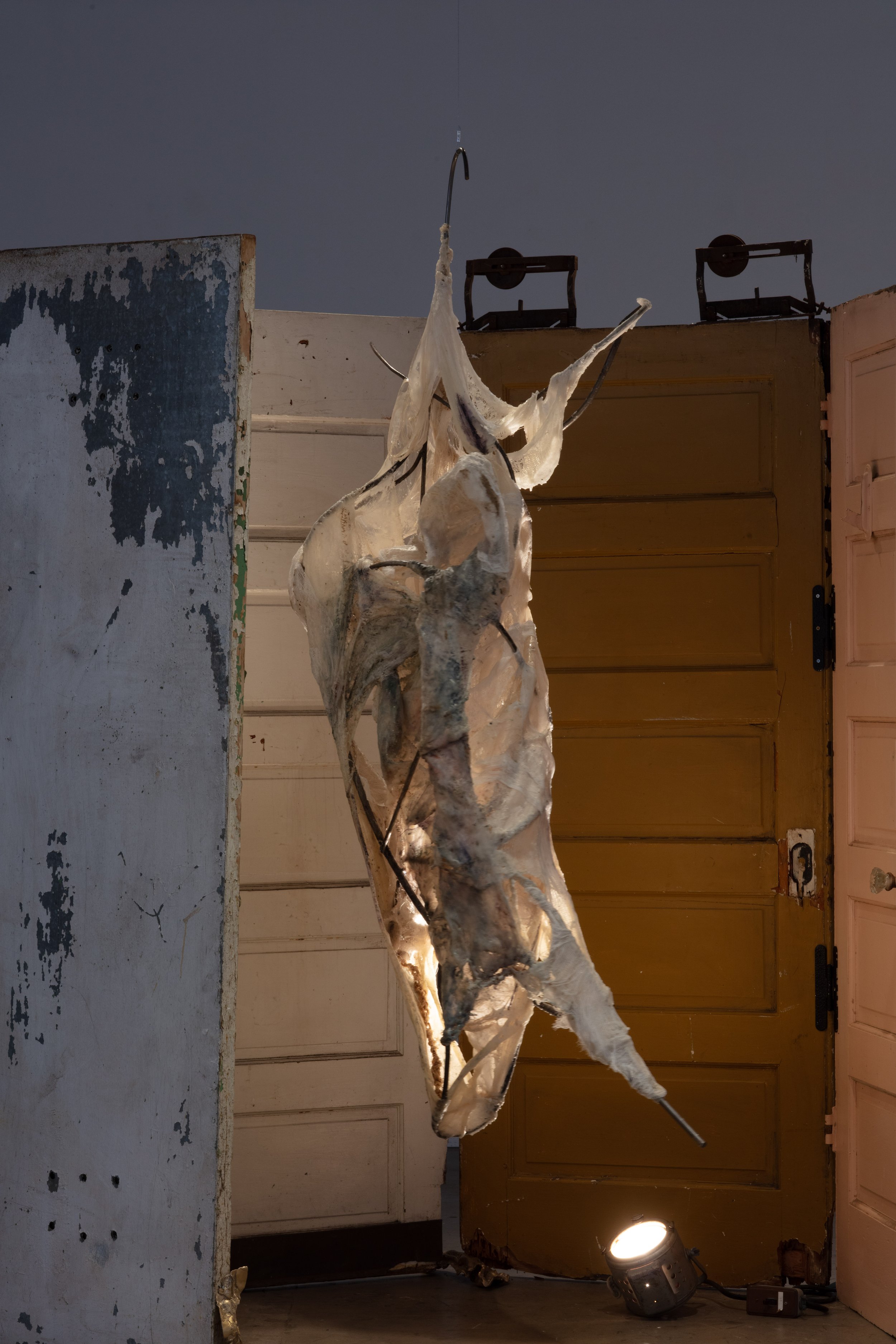 A ghostly sculpture of a creature with elongated limbs, skeletal features, and a ribcage, suspended on a metal hook in an indoor setting with wooden doors and stage lights ceiling.