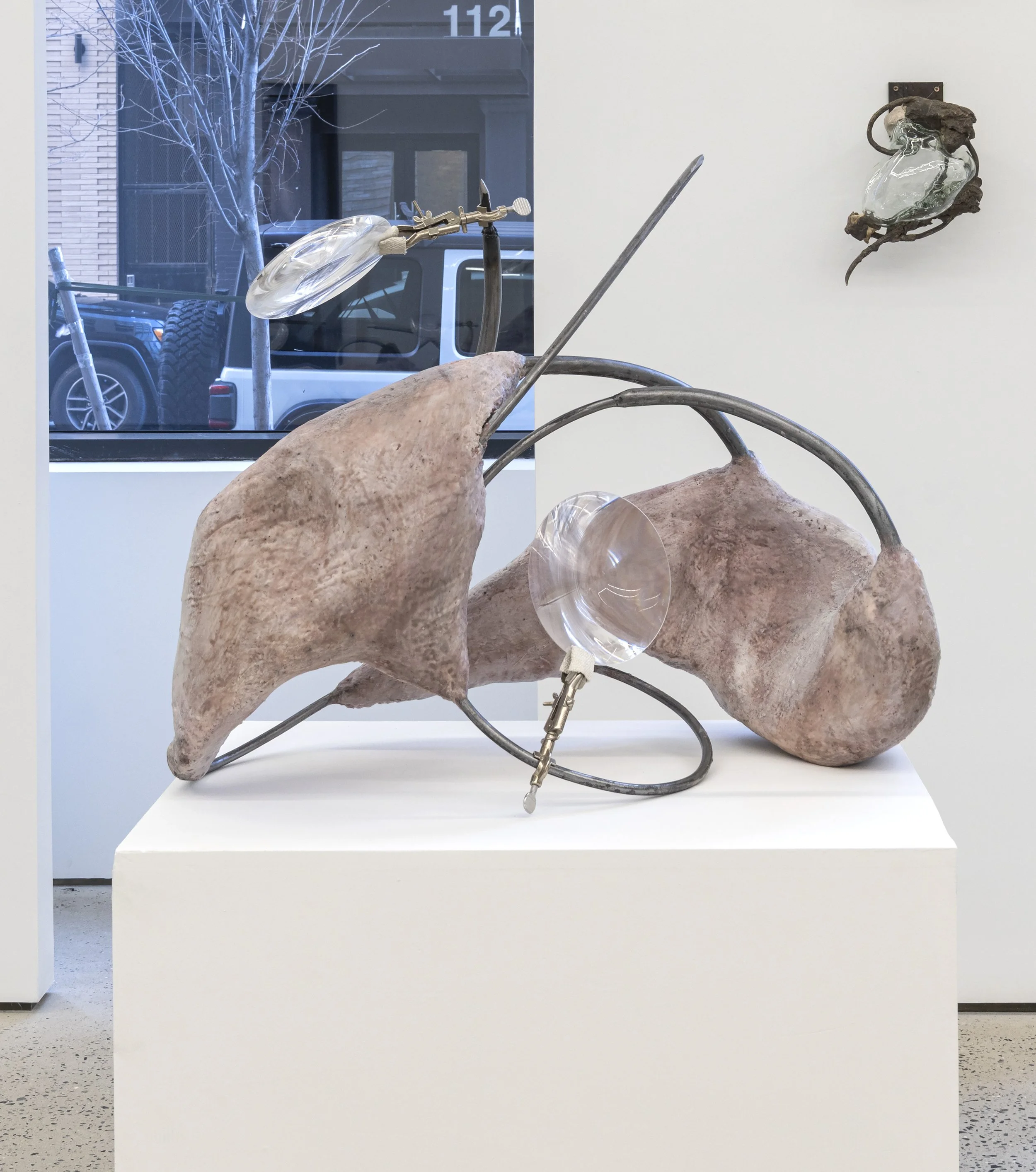 A mixed-media art sculpture features a rock and metal elements, with clear glass or acrylic pieces attached to metal wires and a clamp, positioned on a white pedestal inside an art gallery.