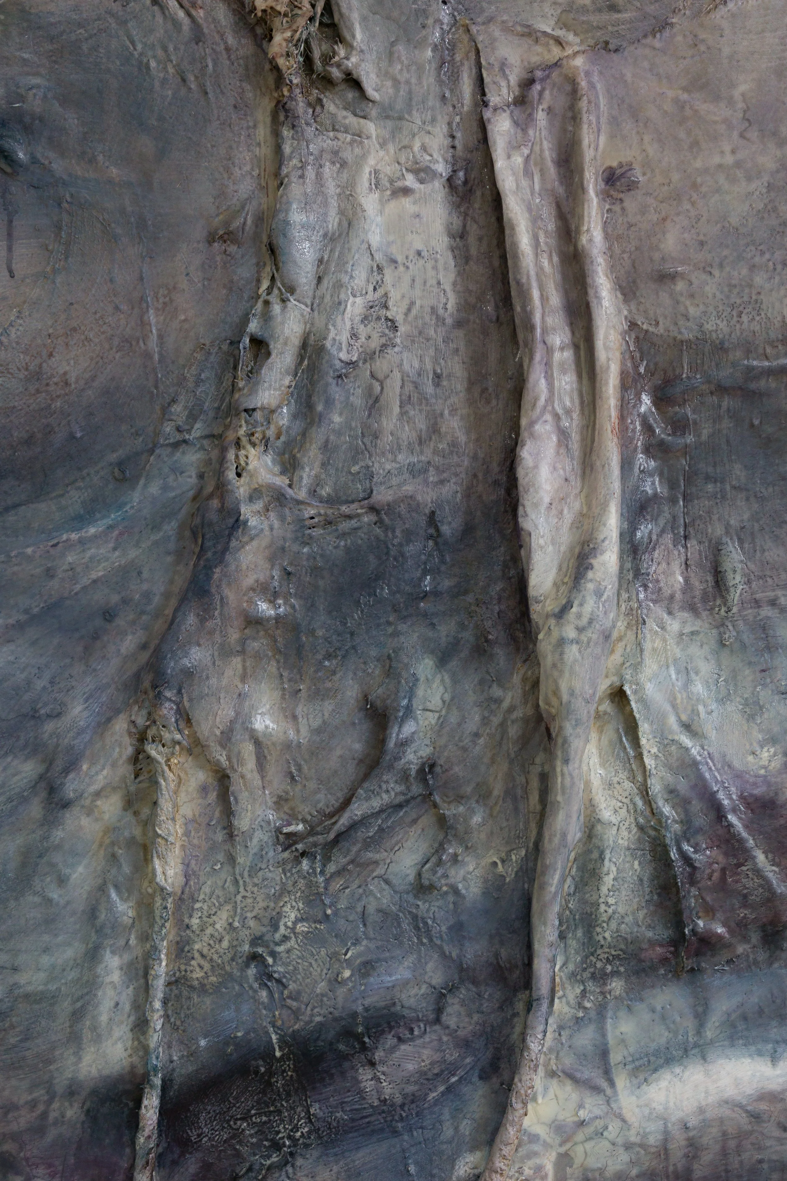Close-up of a textured, rocky surface of a painting with various shades of gray, brown, and hints of purple, showing encaustic formations. Looks like a close-up of a fossilized skeleton embedded in rock, with bones and structure clearly visible.