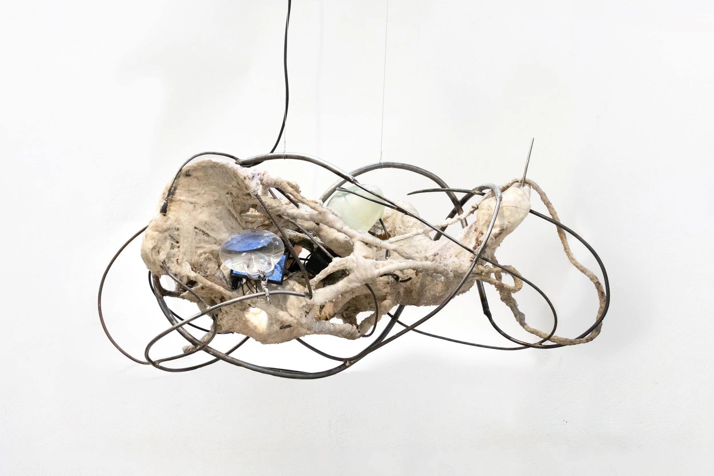 An art installation resembling a vintage car is constructed with irregular, bone-like materials and metal wires, with small glass and plastic objects embedded within.