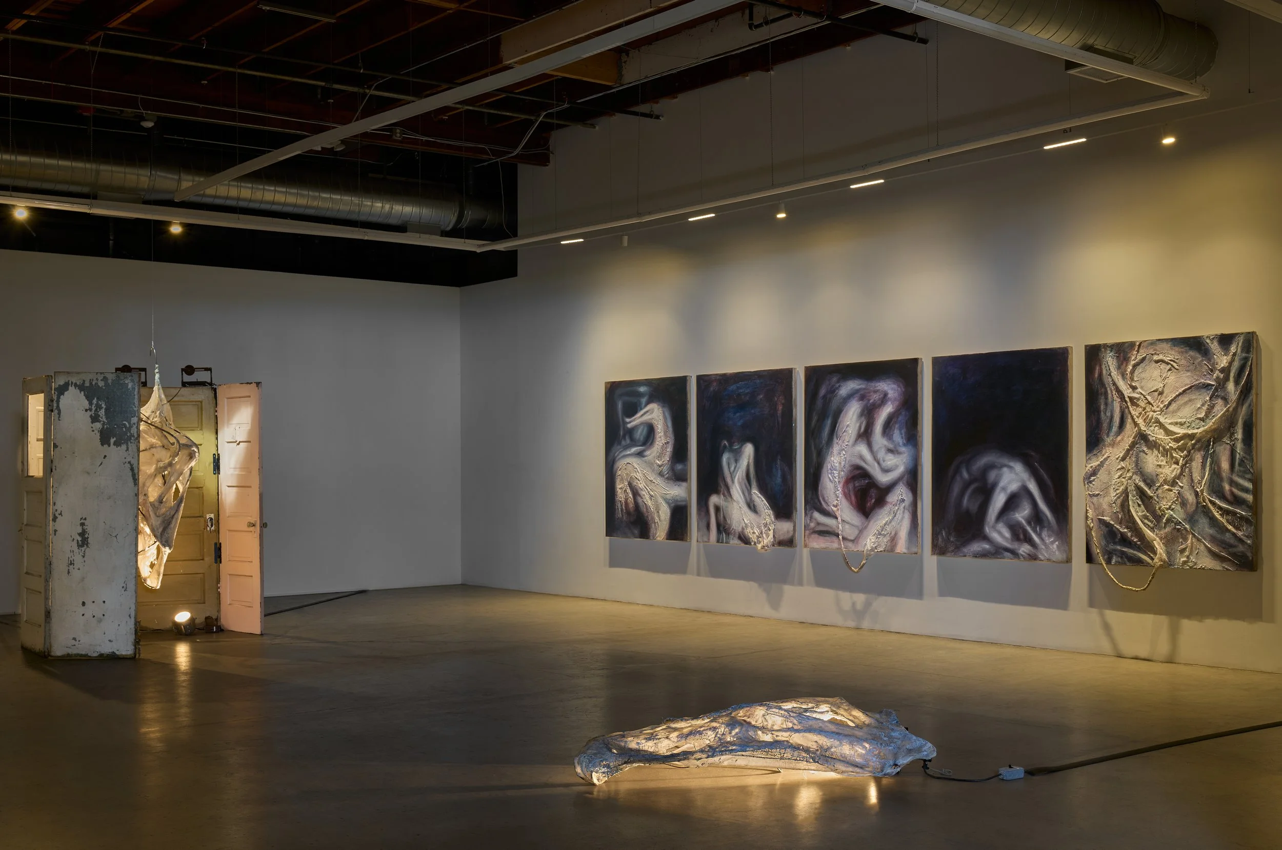 Gallery with abstract art paintings on a white wall and an installation piece on the floor, with a partly open door, in a modern art space.