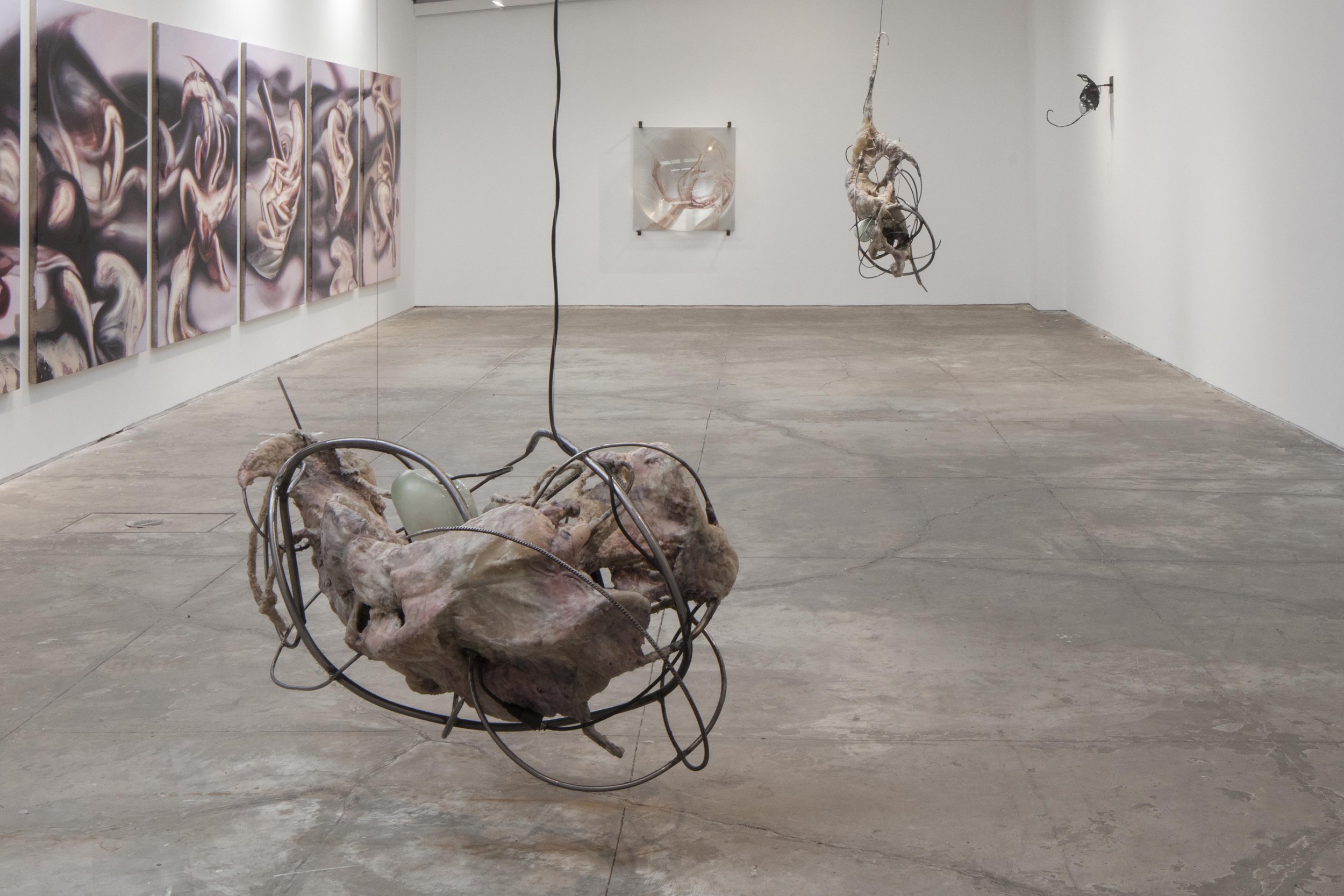 Art installation featuring a sculpture of an abstract, organic shape made from intertwined materials and metal wires, suspended from the ceiling, in an art gallery with white walls and concrete floor, alongside a multi-panel abstract painting on the wall.