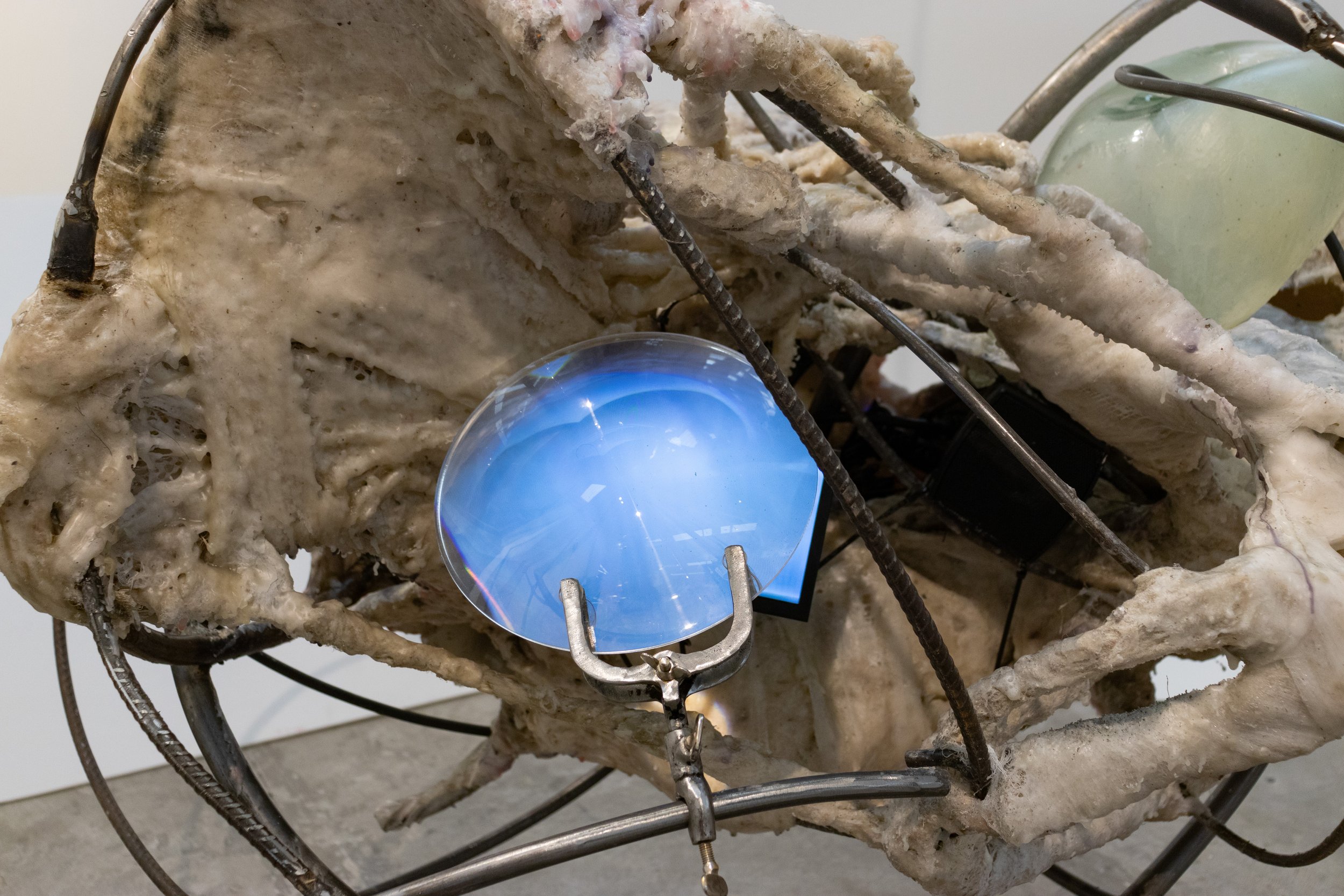 Mixed media art installation featuring a complex structure of intertwined bones, wires, and electronic components, including a lit blue LED light.