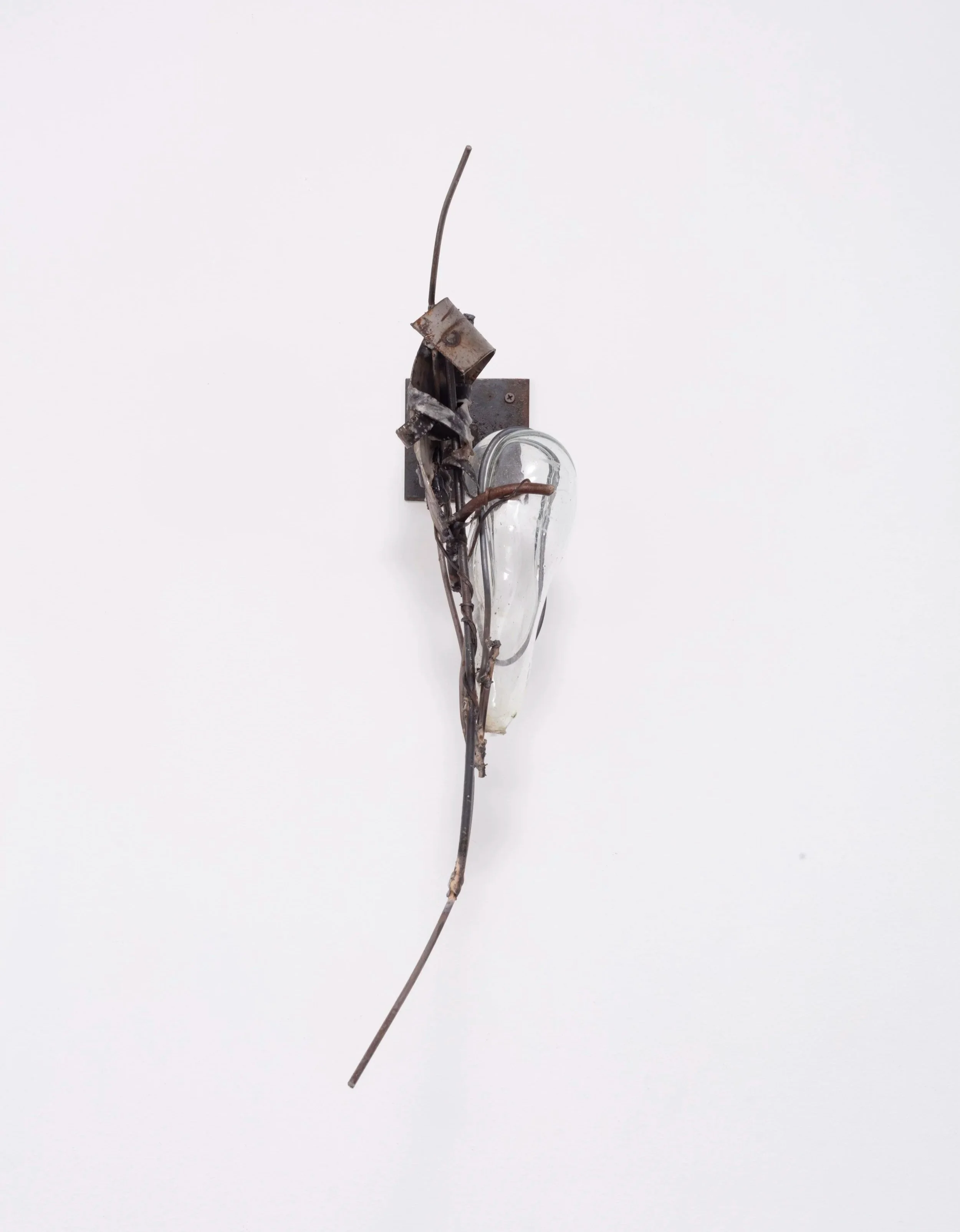 An art sculpture composed of a light bulb, metal parts, and a long wire. The metal parts are mounted on a white wall, with a plain background.