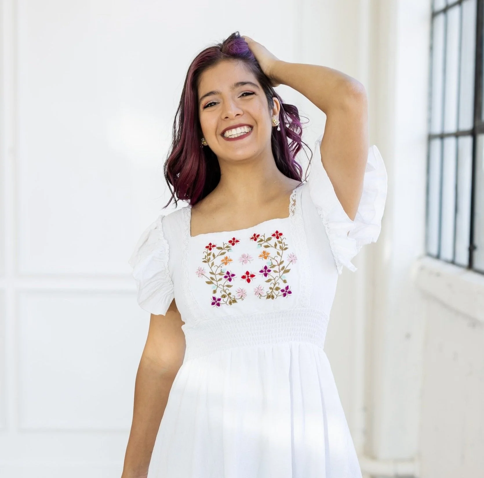 Jimena Montserrat, smiling and wearing in a white dress with floral embroidery, hand touching hair, standing indoors.