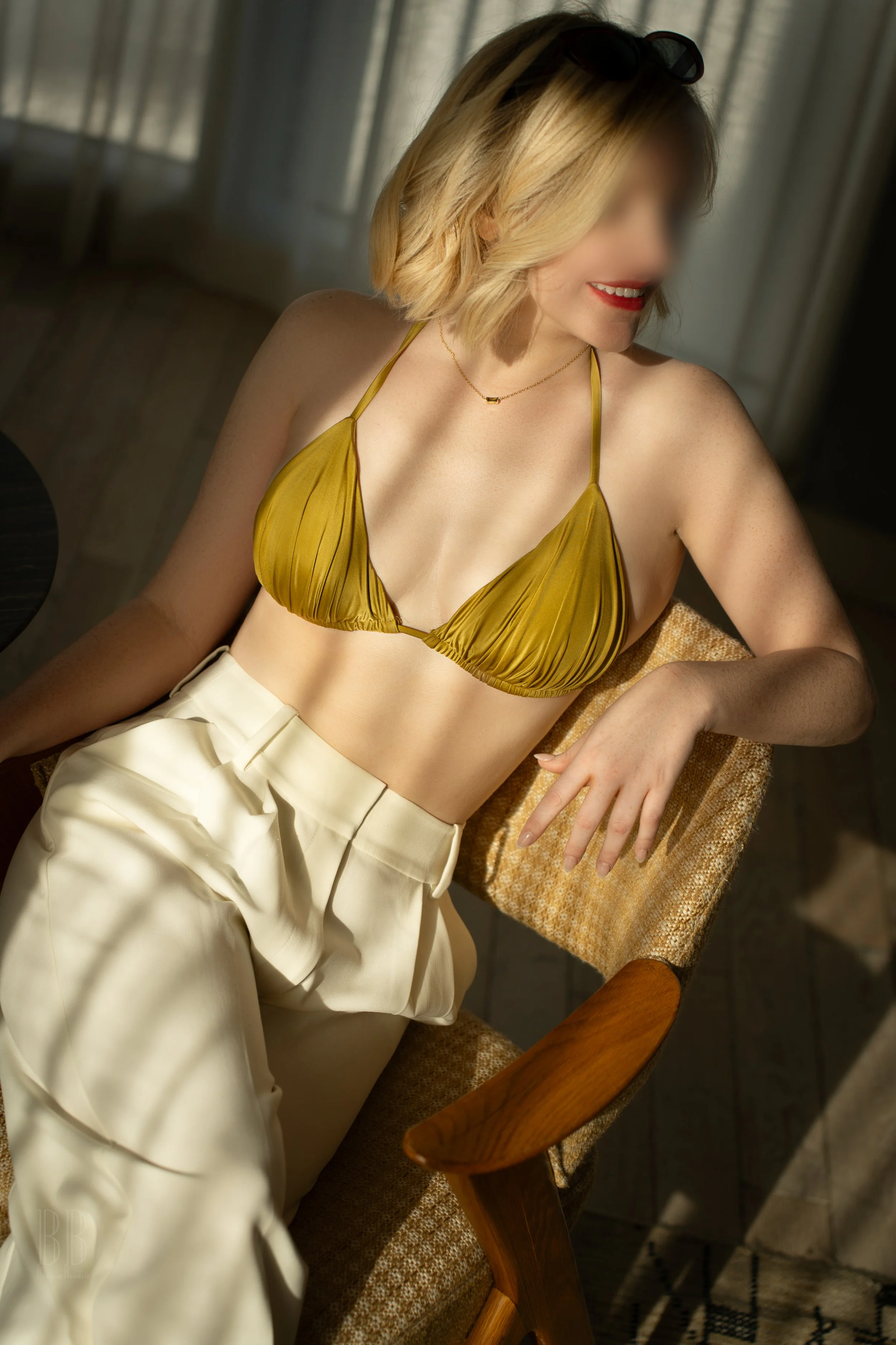 Olive with a blonde bob wearing a yellow bikini top and cream-colored high-waisted pants, sitting on a wooden chair in a sunlit room.