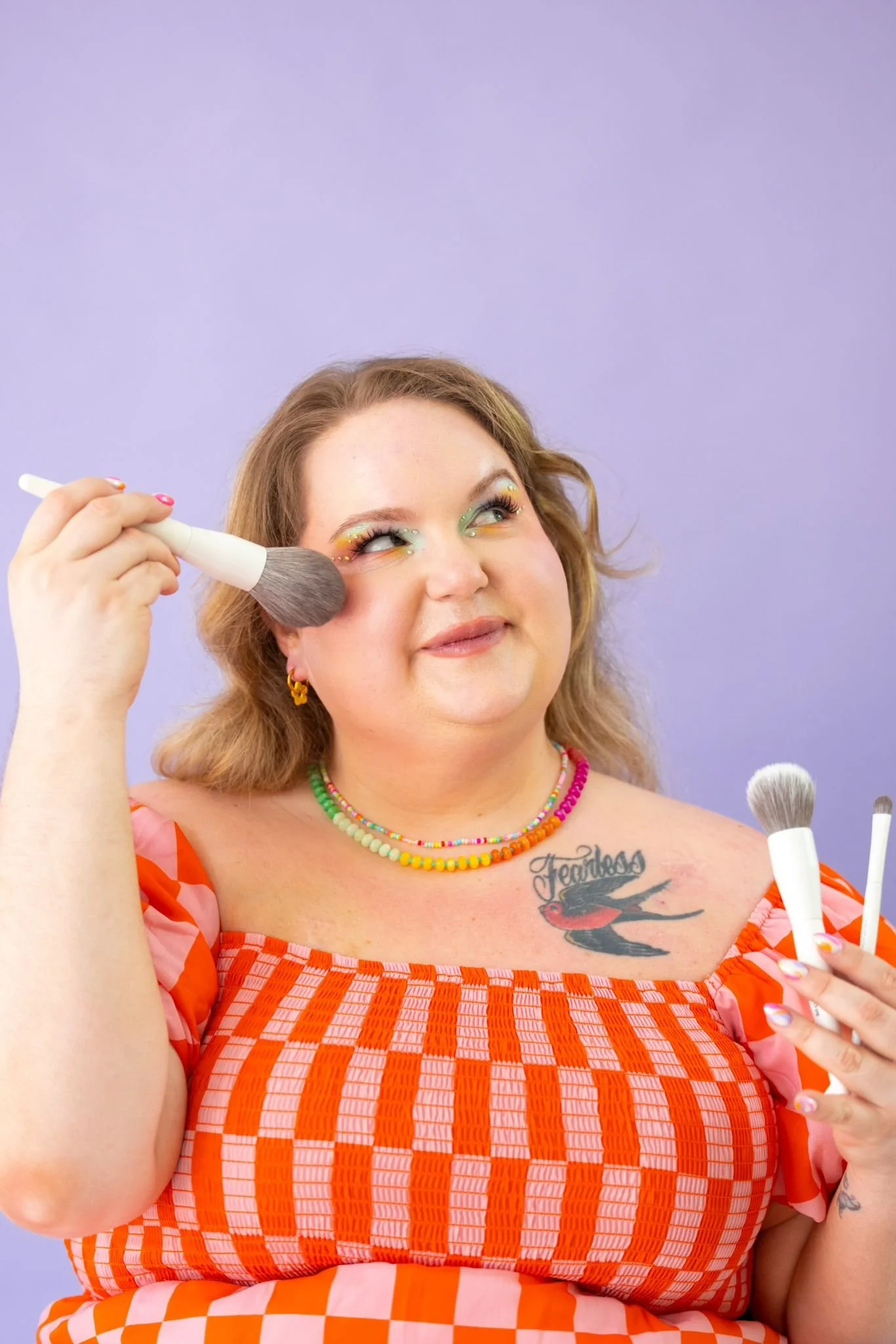 A woman with makeup applying blush with a brush, wearing a colorful checkered dress, rainbow beaded necklaces, earrings, and with a tattoo on her chest that says "Fearless" and features a swallow bird. The background is a plain purple color.