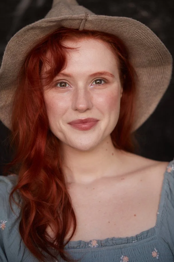 A woman with red hair wearing a wide-brimmed straw hat and a blue off-shoulder top, smiling at the camera against a dark background.