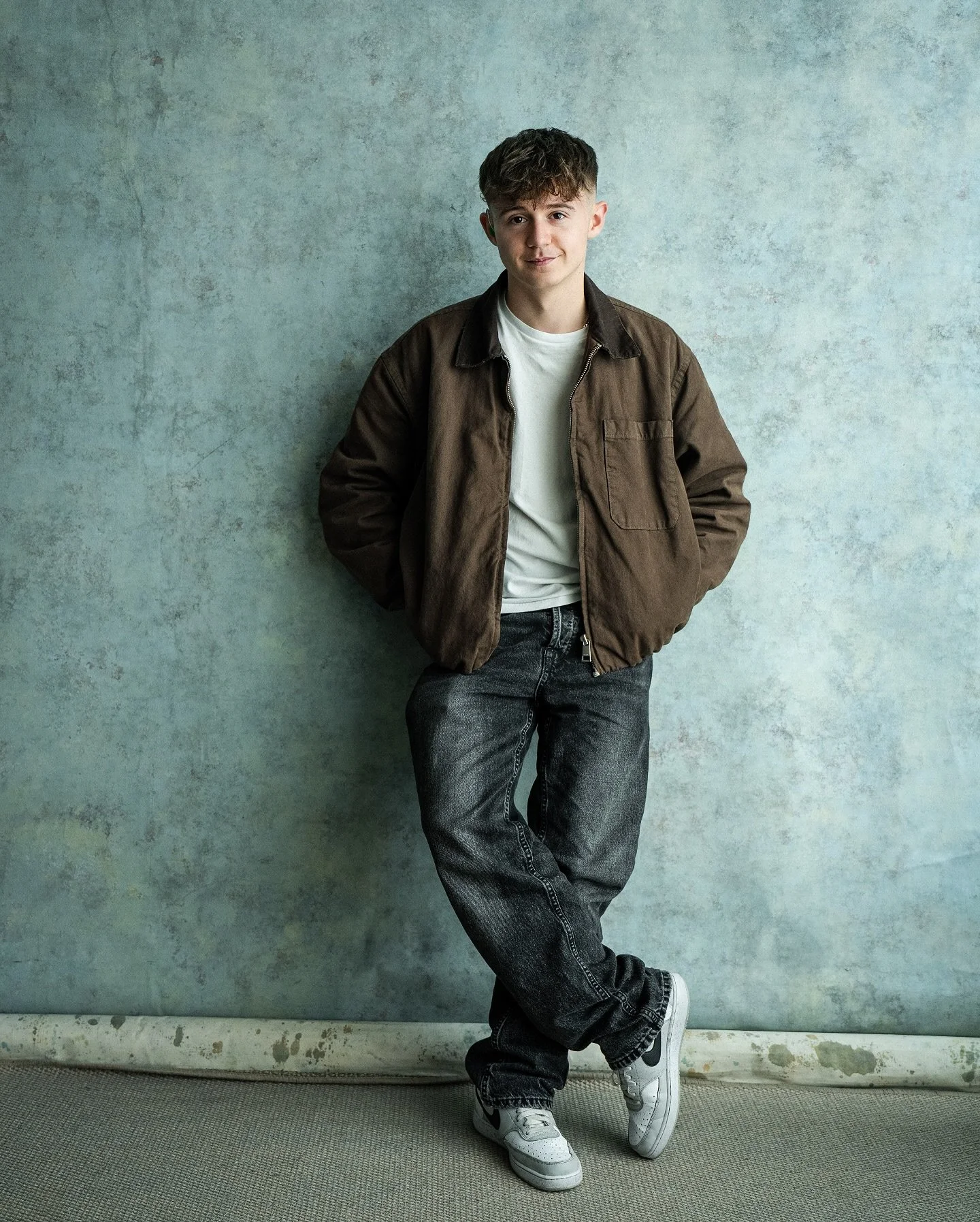 A young man with wavy hair standing against a textured wall, wearing a brown jacket, white T-shirt, black jeans, and white sneakers. He has his hands in his pockets and is smiling slightly.