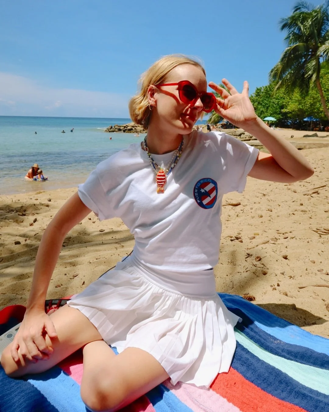 A woman with blond hair wearing red sunglasses, a white t-shirt with a Puerto Rico logo, and a white pleated skirt, sitting on a beach towel on the sand, with palm trees, blue sky, and ocean in the background.
