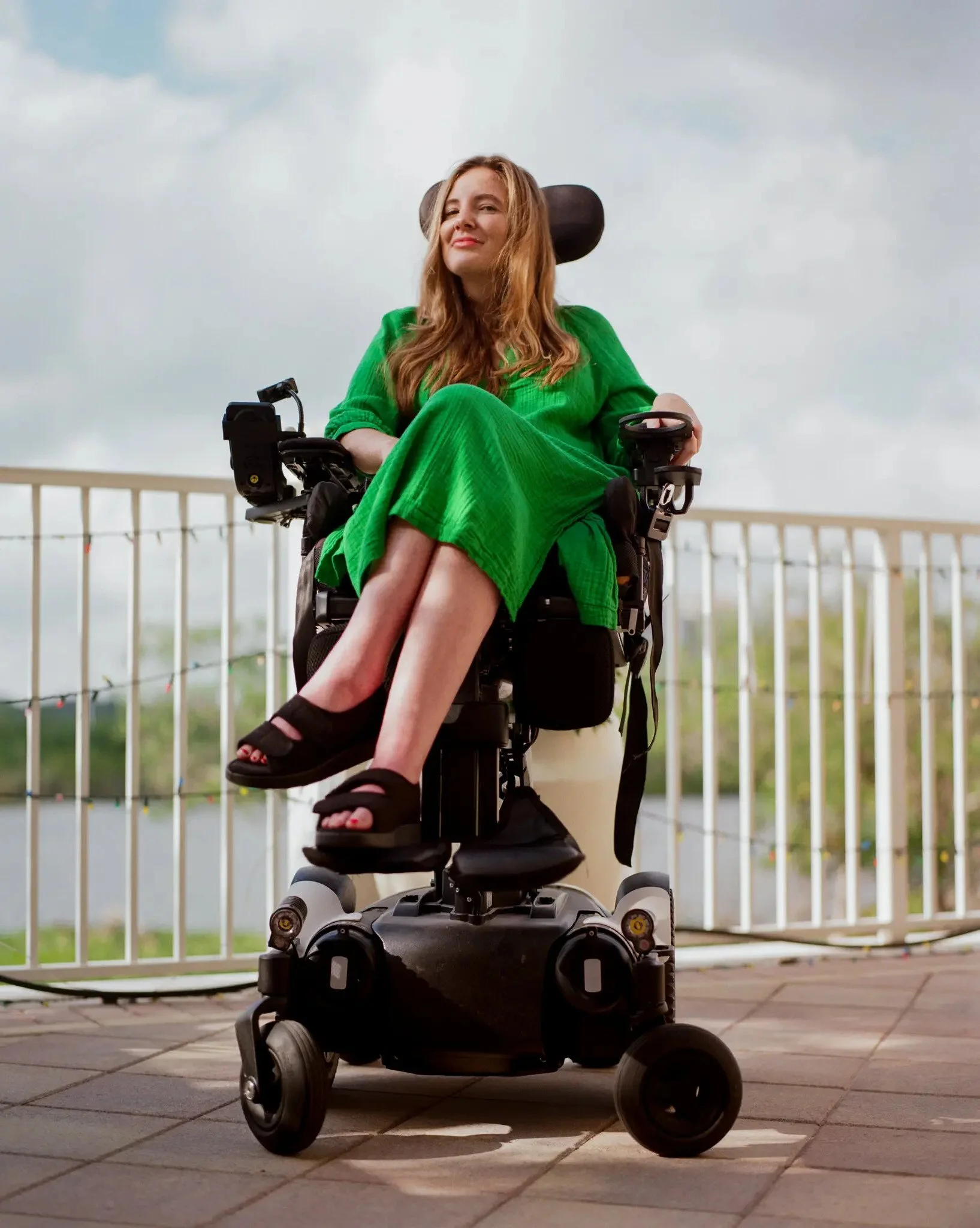 A woman with long red hair sitting in a motorized wheelchair on a patio with tiled flooring, wearing a green dress. She is smiling slightly and looking at the camera, with an overcast sky and a white metal railing in the background.