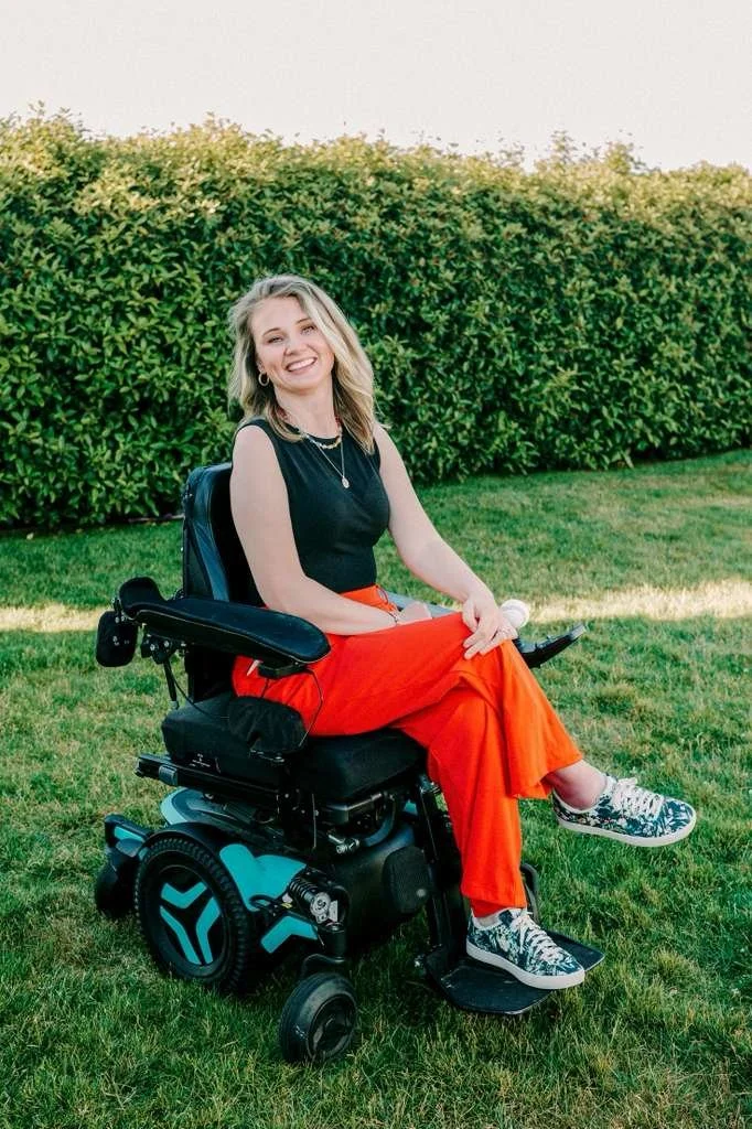 A smiling woman with blonde hair sitting in a powered wheelchair on grass with a green hedge in the background. She is wearing a black sleeveless top, bright orange pants, and patterned sneakers.
