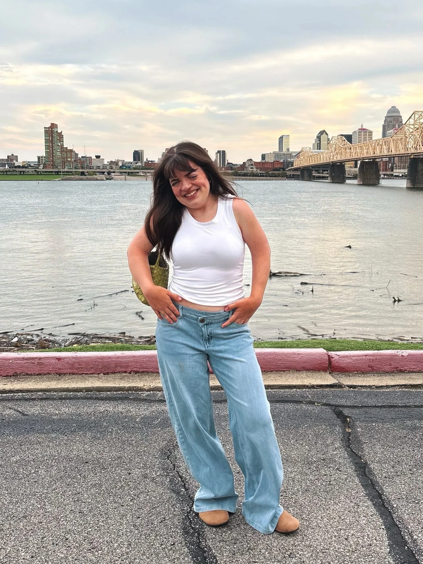 A woman with shoulder-length dark hair smiling and posing with her hands on her hips by a body of water, with a city skyline and bridge in the background during the daytime.