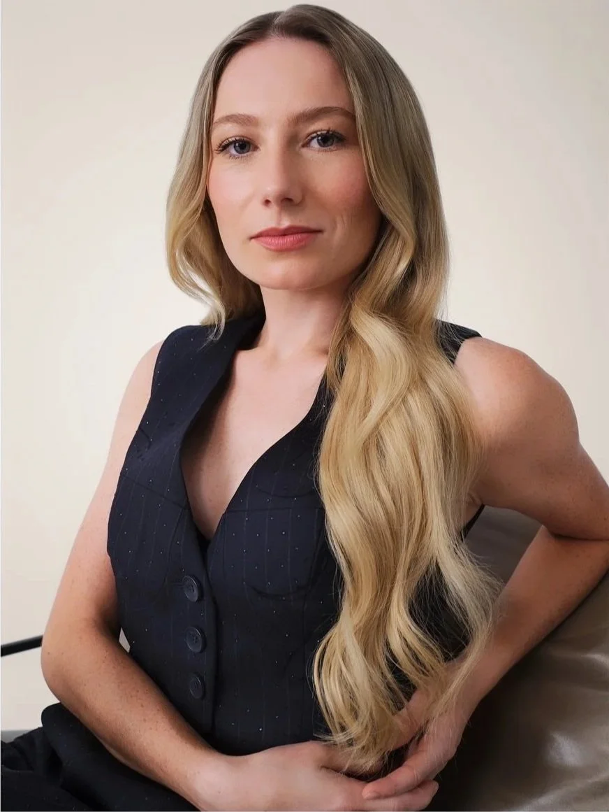 A woman with long, wavy blonde hair in a black pinstripe sleeveless vest, sitting with her arm resting on her lap, looking at the camera against a plain light background.